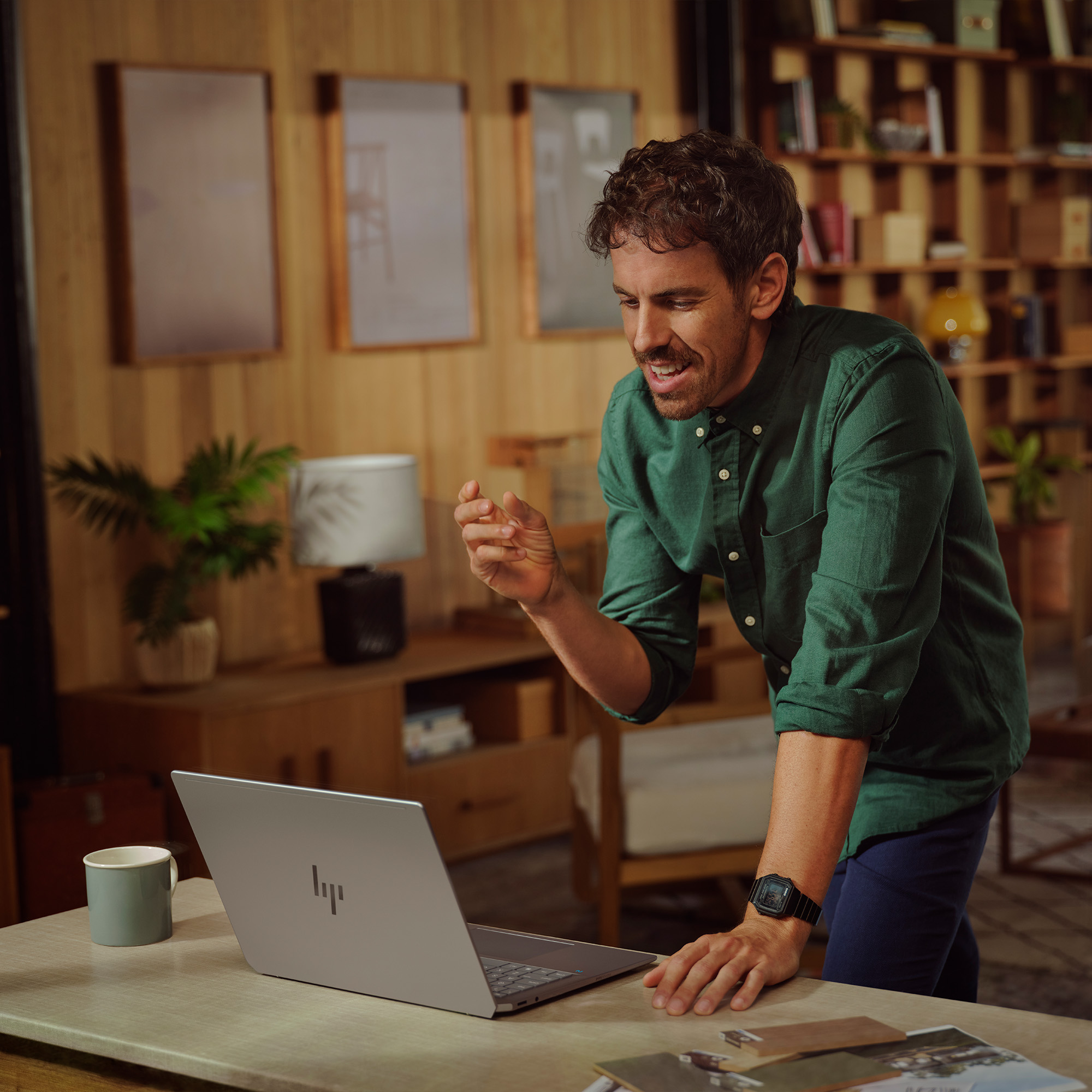 Mężczyzna w zielonej koszuli gestykuluje, patrząc na laptopa. Kubek i czasopisma leżą na biurku. Tłem jest drewniana ściana.