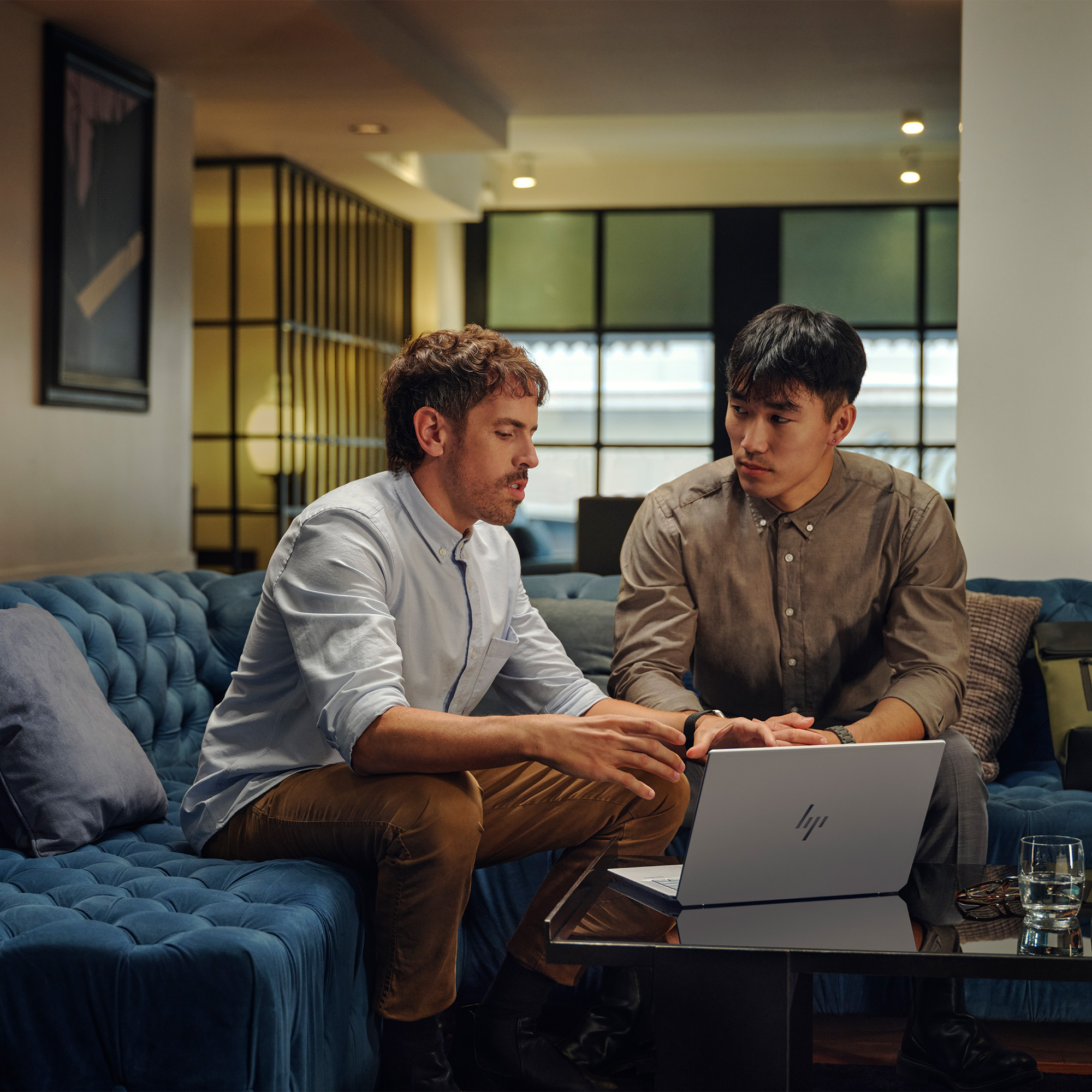 Dwóch mężczyzn siedzi na niebieskiej sofie, patrząc na laptopa. Jeden gestykuluje, drugi obserwuje. Szklanka wody na stole.