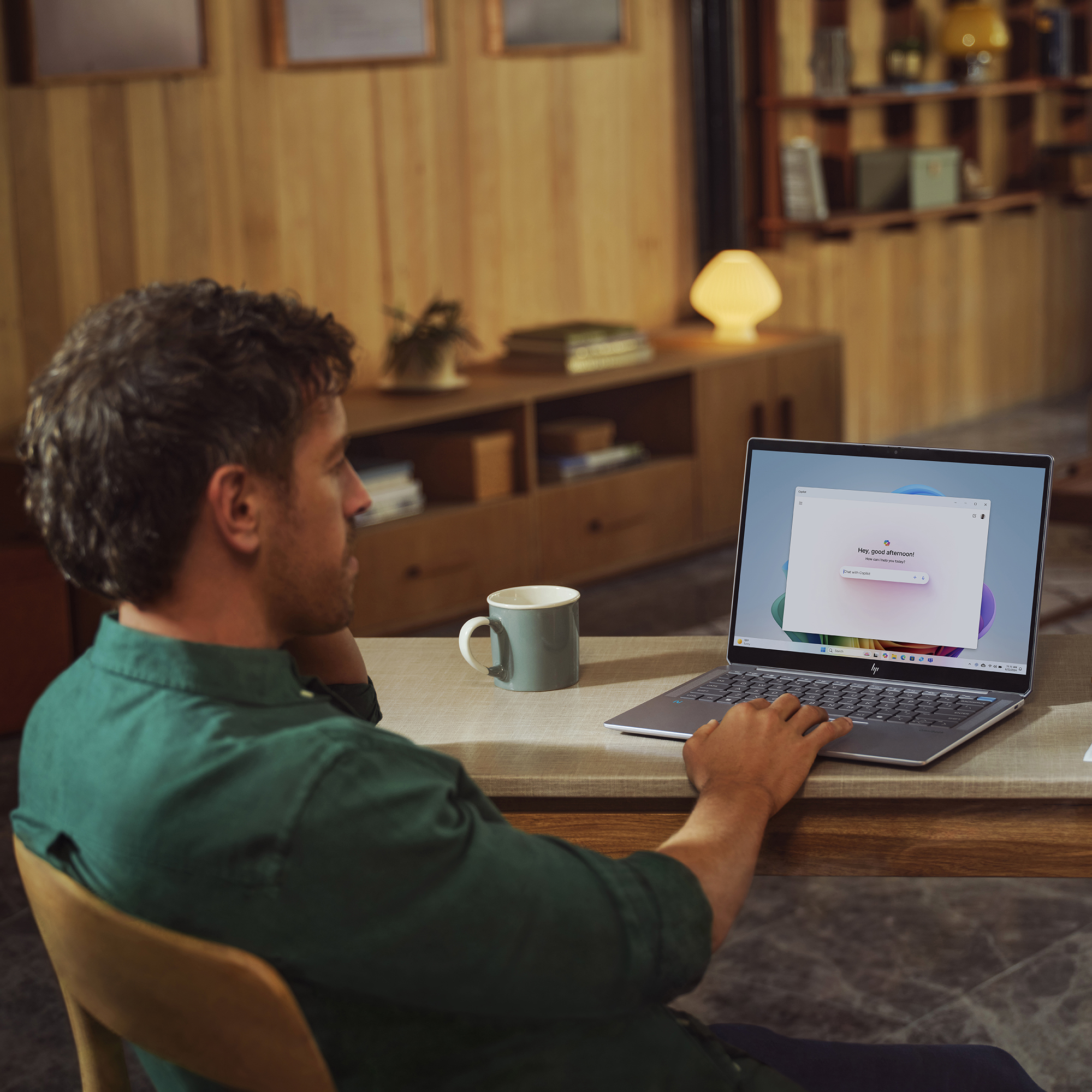 Mężczyzna używa laptopa, siedząc przy drewnianym stole z kubkiem. Ekran laptopa pokazuje interfejs czatu. Drewniana ściana w tle.