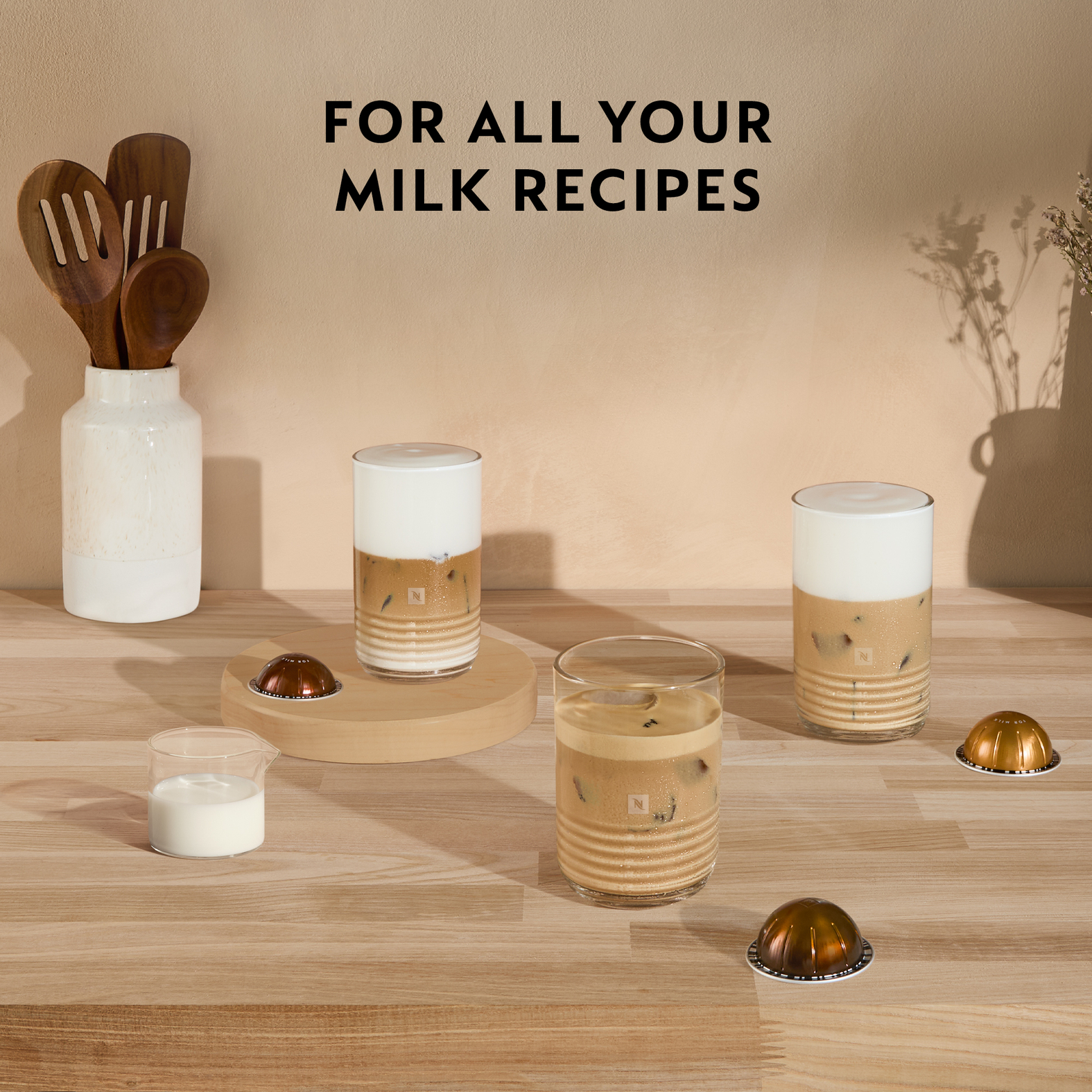 Kaffeegetränke mit Milch werden zusammen mit Kaffeekapseln und Milch angezeigt. Holzlöffel und eine Vase sind im Hintergrund zu sehen.