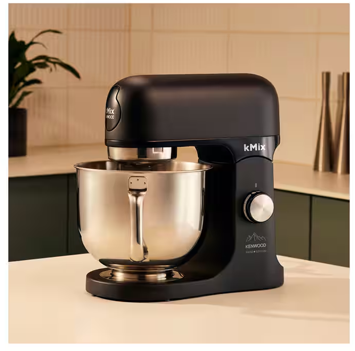 Schwarzer Kenwood kMix Küchenmixer mit Edelstahlschüssel auf weißer Oberfläche. Der Hintergrund zeigt eine Küchenszene.