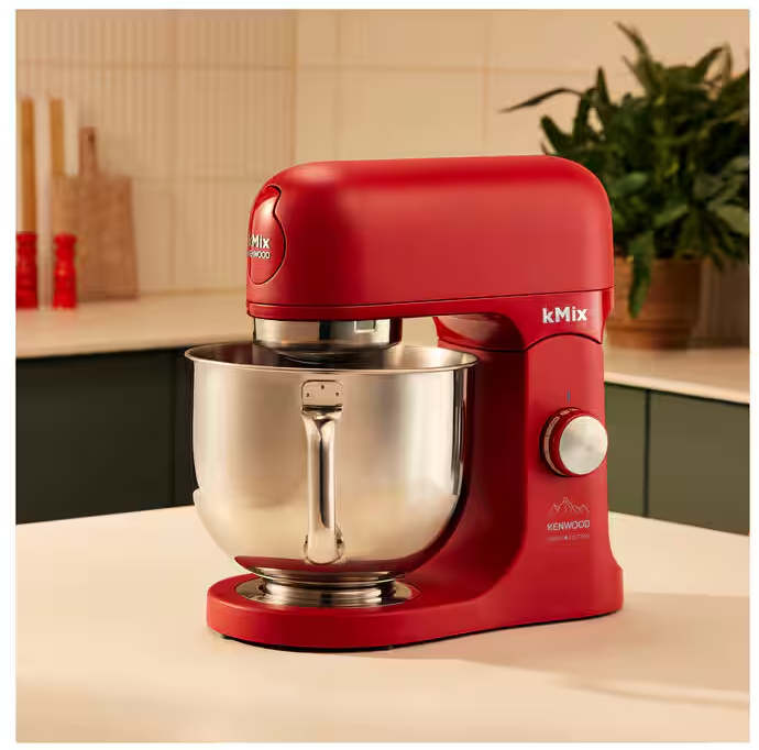 Ein roter Kenwood kMix-Küchenmixer mit einer Edelstahlschüssel steht auf einer weißen Oberfläche. Der Hintergrund umfasst eine Küche und grüne Pflanzen.