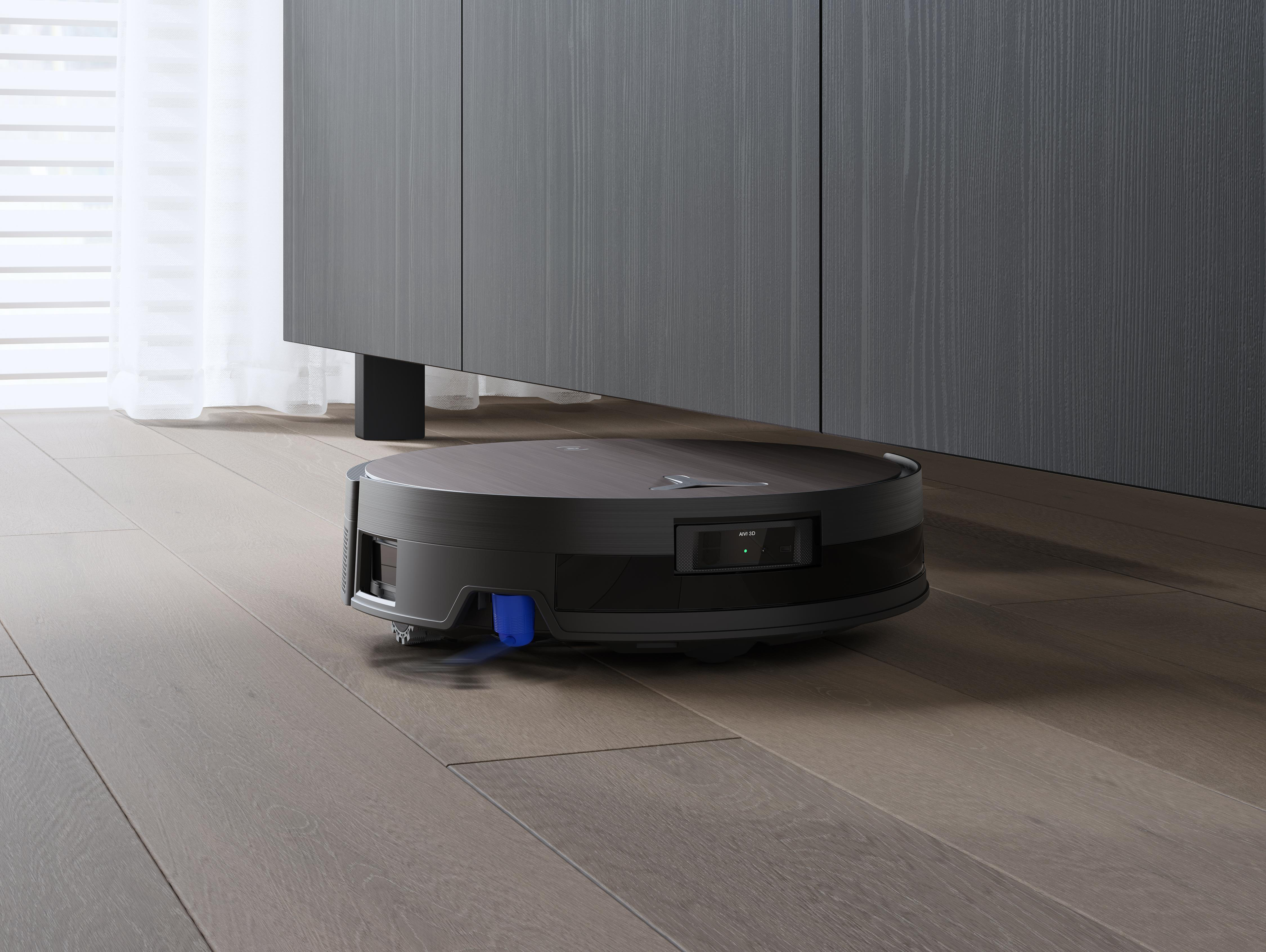 Ein schwarzer Roboter-Staubsauger steht auf einem Holzboden unter einem dunklen Schrank. Fenster und Vorhänge sind im Hintergrund.