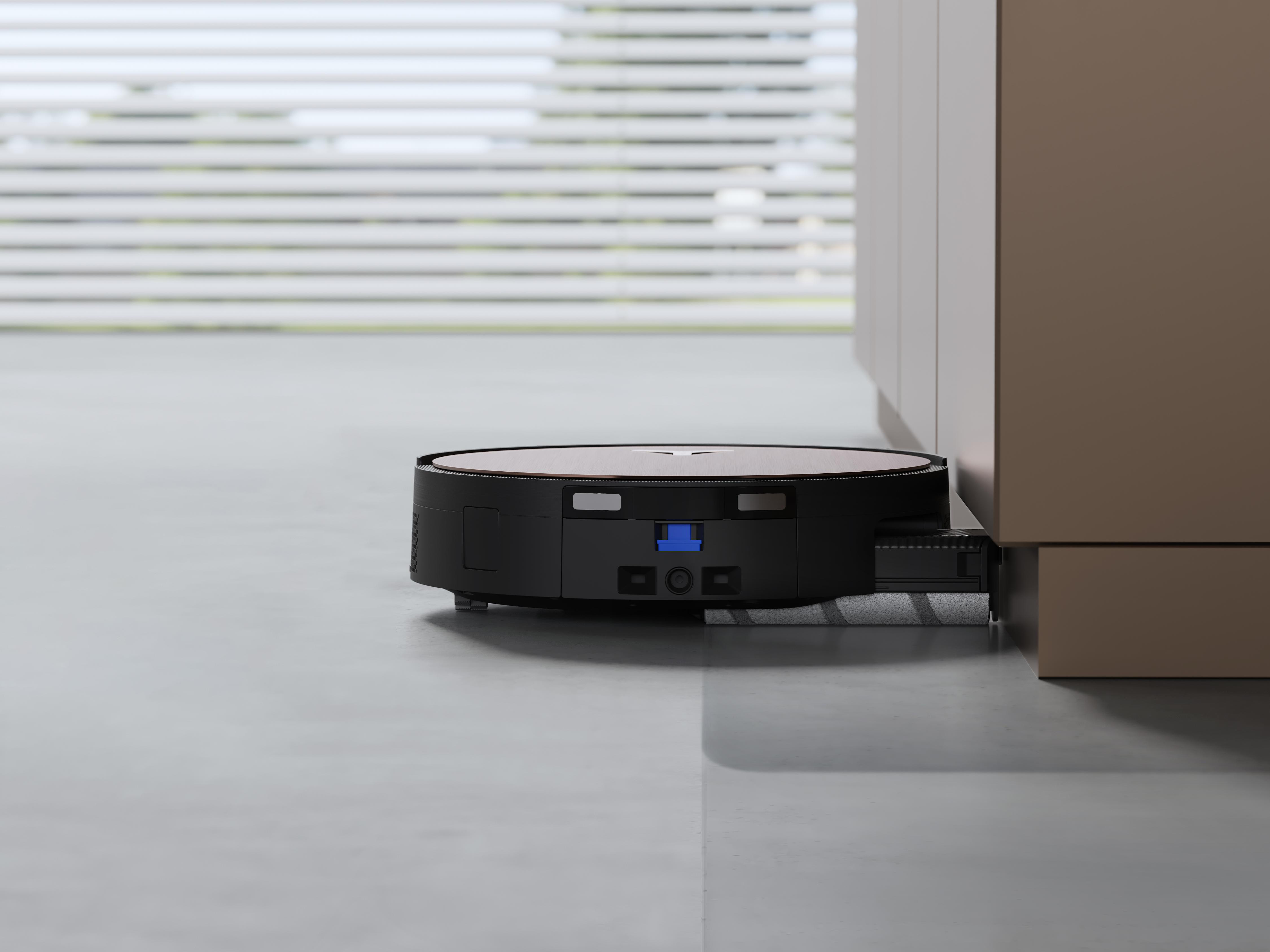 Ein schwarzer Roboter-Staubsauger auf grauem Boden. Er befindet sich in der Nähe eines braunen Schranks. Im Hintergrund befinden sich Jalousien.