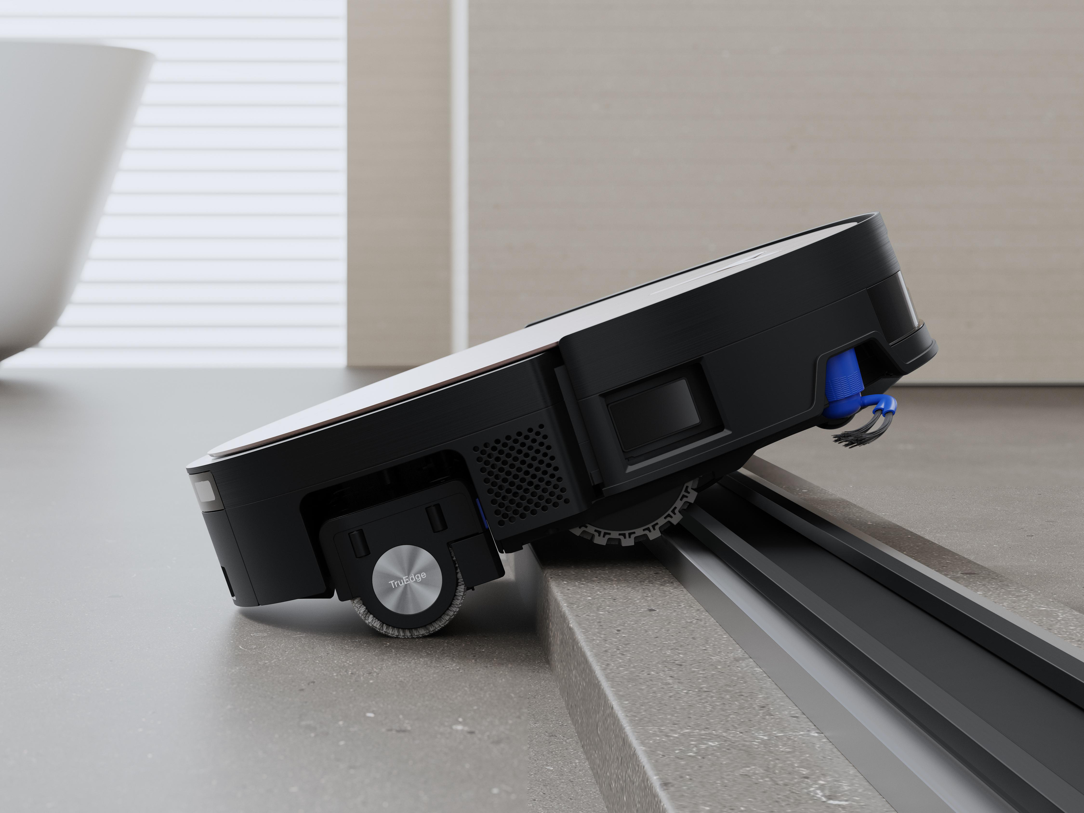 Ein schwarzer und silberner Roboter-Staubsauger auf grauer Oberfläche, der sich teilweise eine Stufe hinunterbewegt. Blaue Bürste und weiße Wand im Hintergrund.