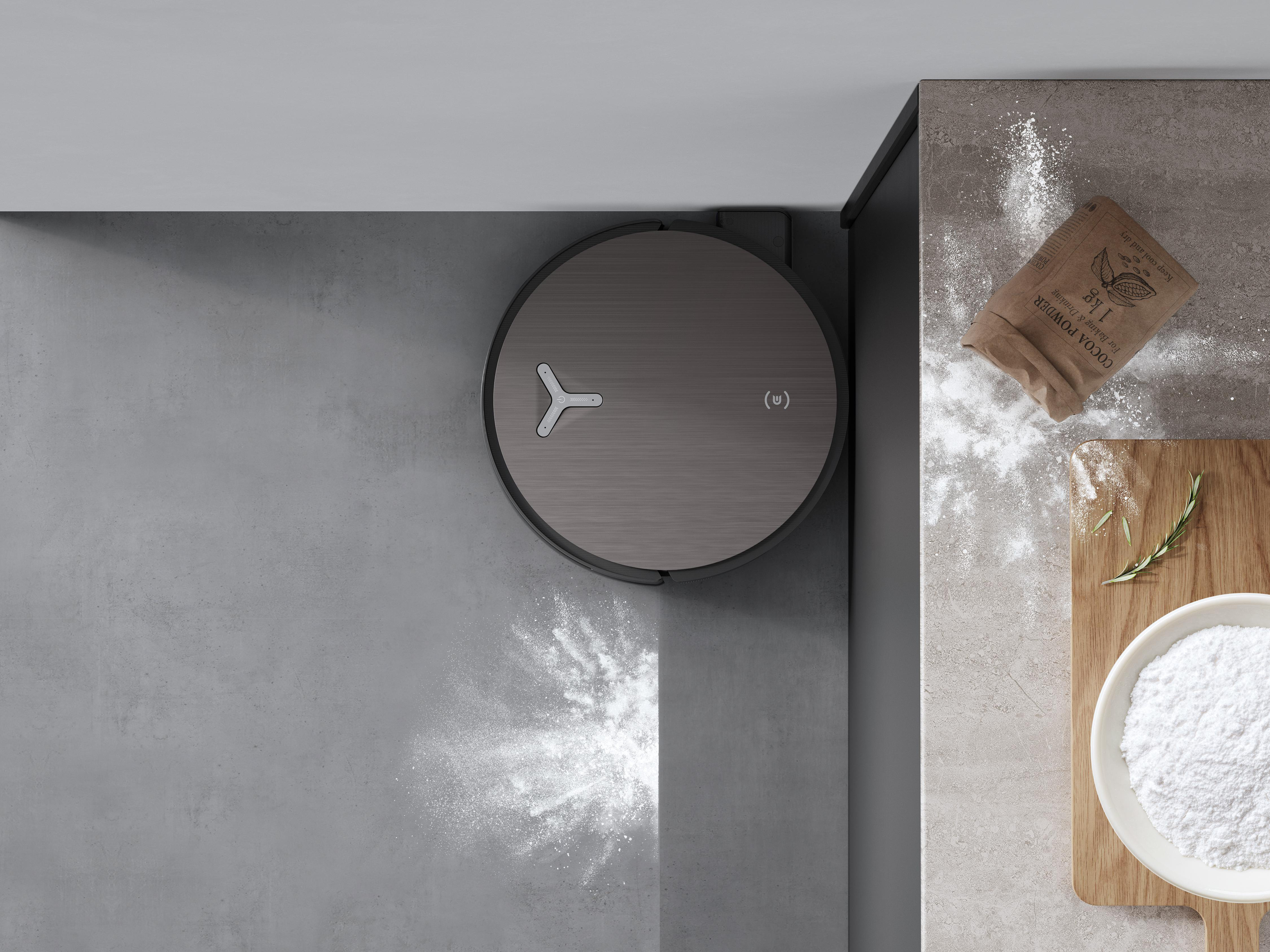 Ein Roboter-Staubsauger steht auf einem grauen Boden, mit verschüttetem Kakaopulver und Backzutaten. Ein Holzbrett mit einer Schüssel Mehl.