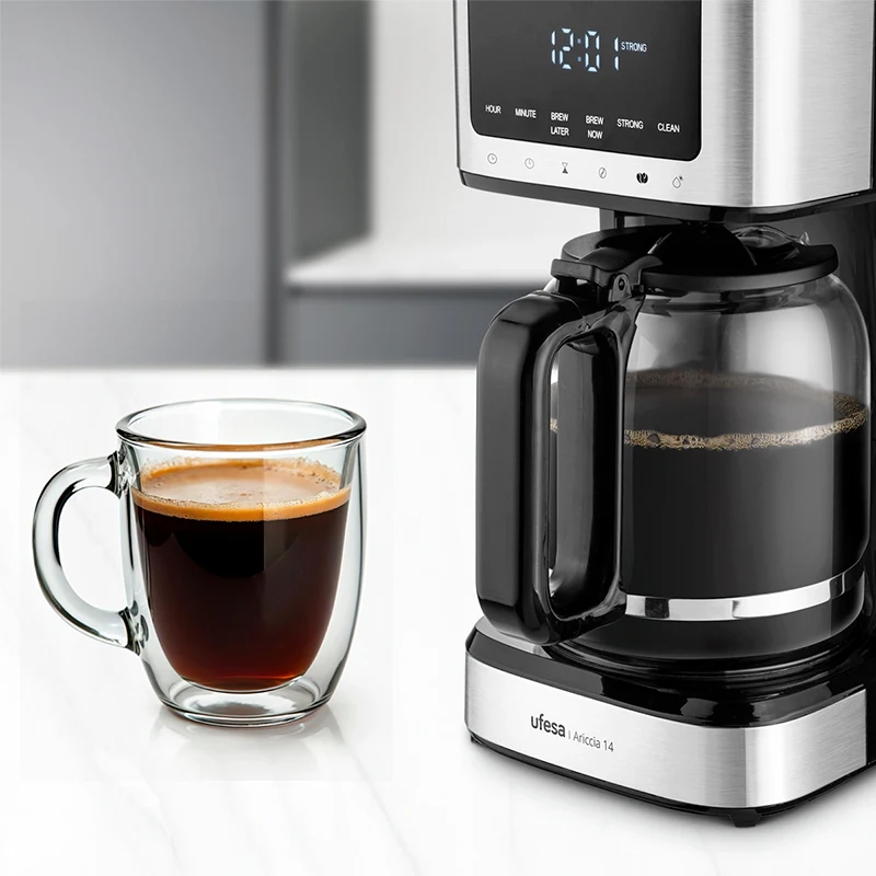 Una cafetera con una jarra de vidrio y una taza de café. La máquina tiene una pantalla digital. La taza es de doble pared.
