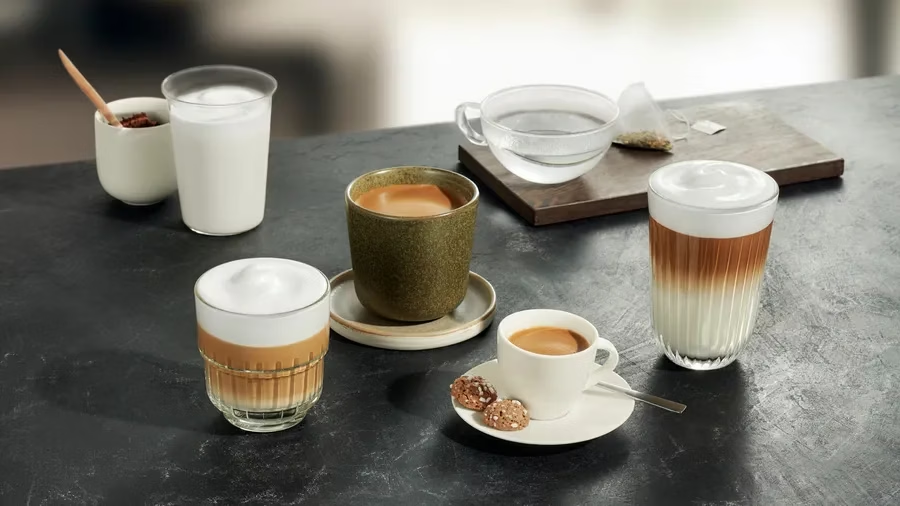 Több kávéital látható egy sötét felületen. Lattékat, eszpresszókat és egy csésze vizet tartalmaz egy teafilterrel.