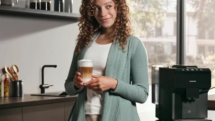 Nő egy lattét tart a konyhában. Zöld kardigánt visel. Jobbra kávéfőző. Konyhai háttér.