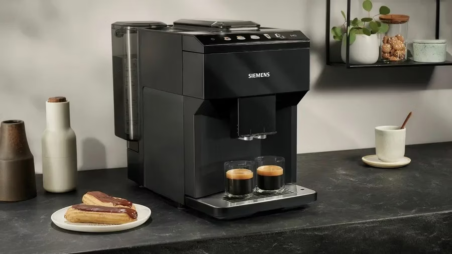 Egy fekete Siemens eszpresszó gép kávét főz két pohárba. Ekler fánkok vannak egy tányéron. Egy csésze és egy polc üvegekkel láthatók.