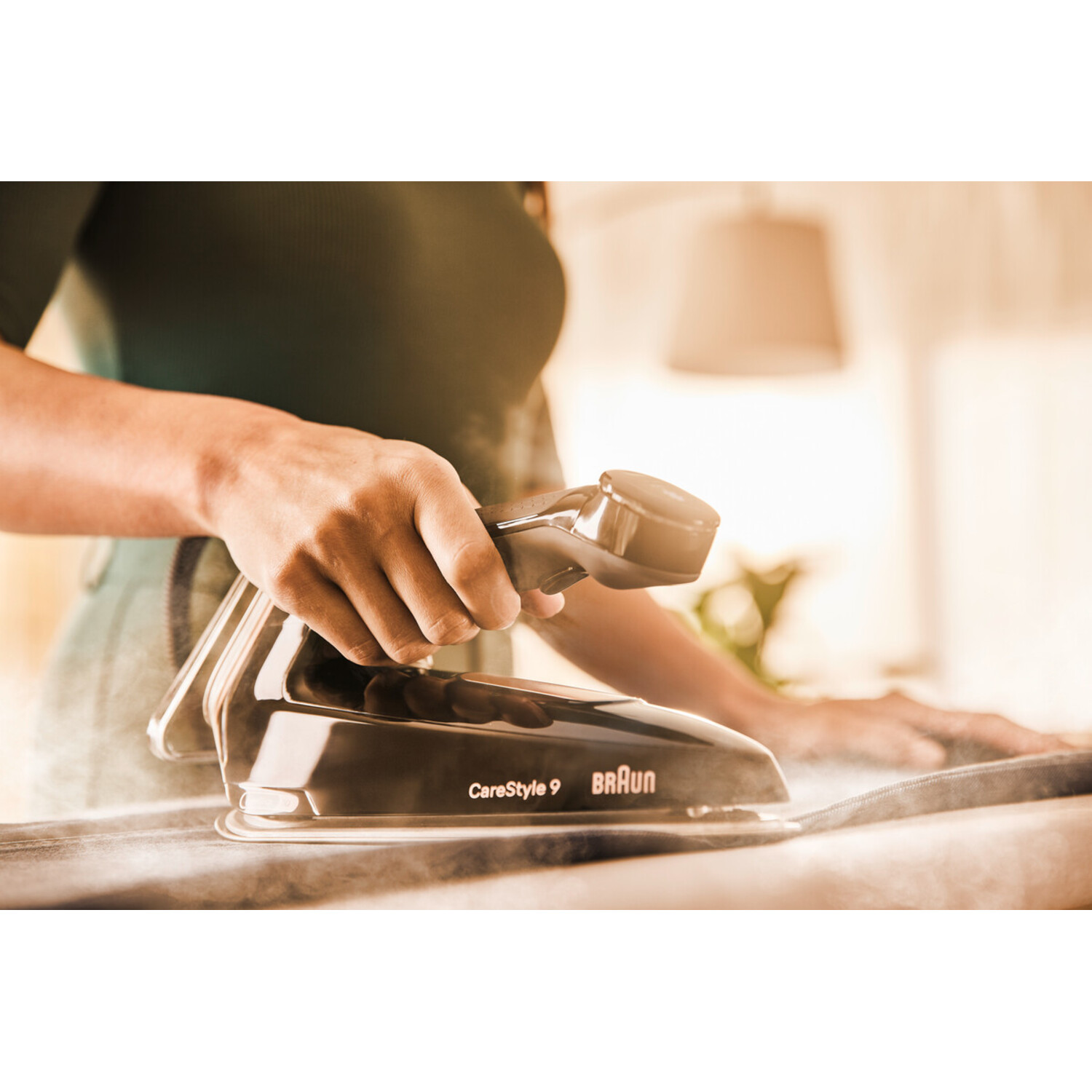 A person ironing clothes with a black iron. Steam rises. The iron is on a white ironing board. Braun CareStyle 9 is on the iron.