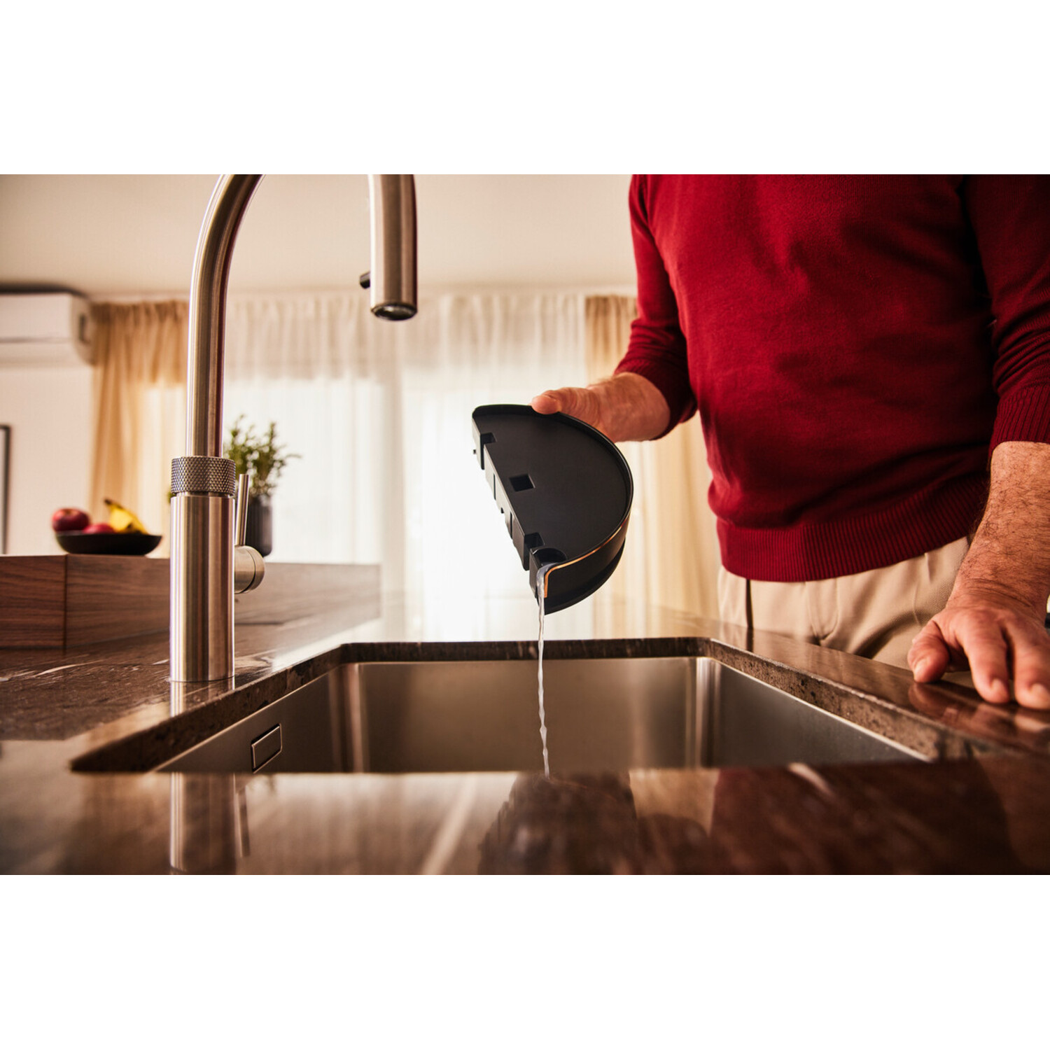 A person in a red sweater empties a black container into a stainless steel sink. A faucet is in the background.