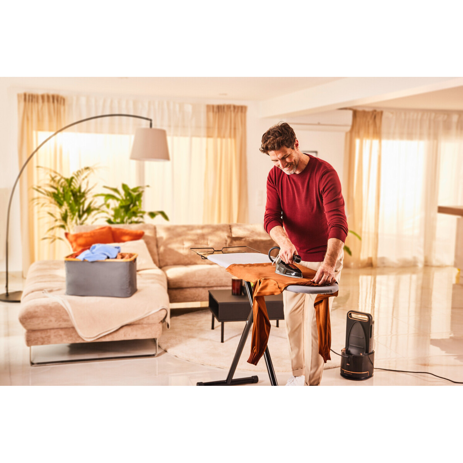A man irons pants on an ironing board. There's a sofa, basket of clothes, and a steam generator. The room has natural light.