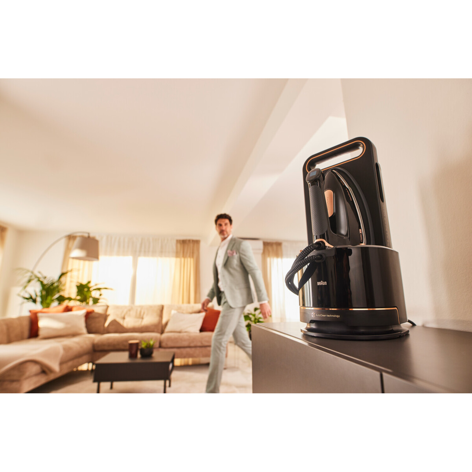 A man walks past an iron on a stand. The iron is black with gold accents. A sofa, plants, and a lamp are in the background.