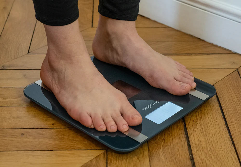 Pieds nus sur une balance numérique. L'écran affiche un poids. Le sol est en bois.