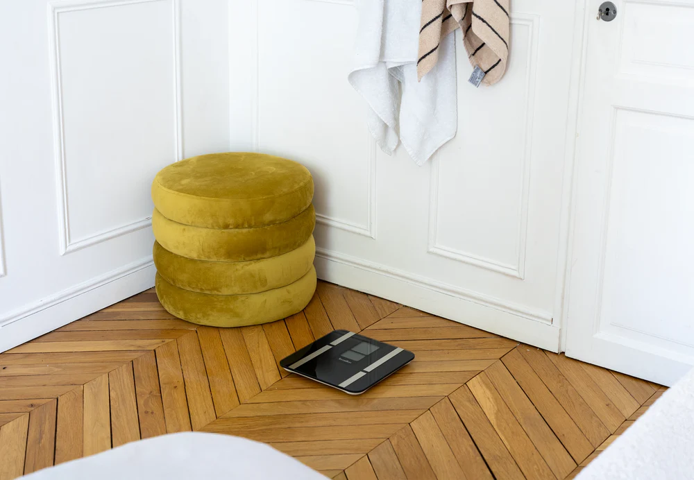 Une scène de salle de bain avec un tabouret jaune, des serviettes et une balance. Plancher en bois, murs blancs et une porte.