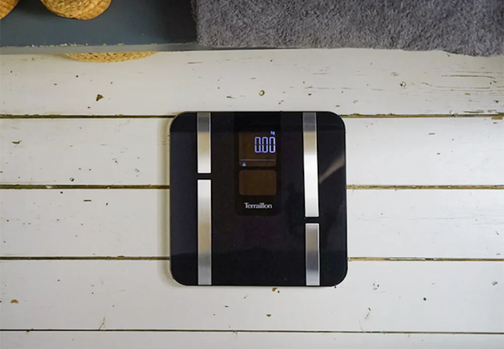 Une balance numérique noire avec une lecture de 0,00 est posée sur des planches de bois blanches. Une serviette grise et un panier sont au fond.