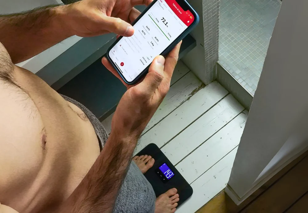 Homme tenant un téléphone avec des données de poids, debout sur une balance. Corps visible, cadre de salle de bain. La balance affiche 14,3, le téléphone affiche 72,1 kg.