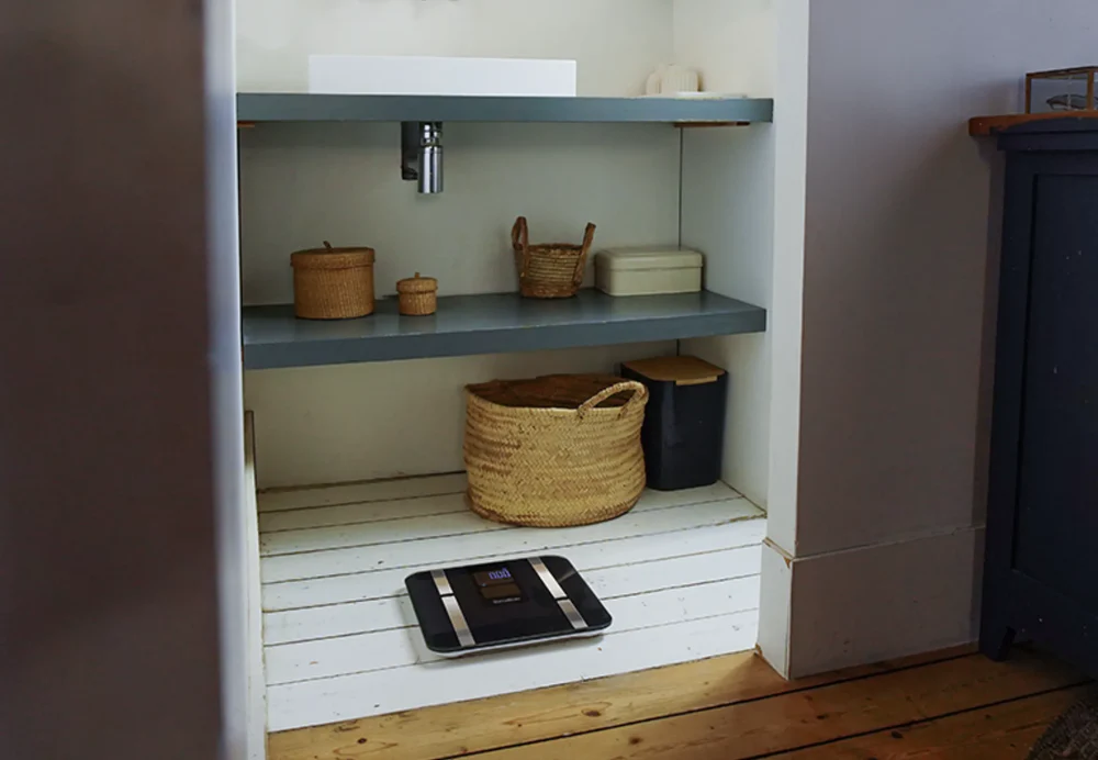 Une salle de bain avec des étagères encastrées. Il y a des paniers, des boîtes et une balance. Un meuble sombre est à droite.
