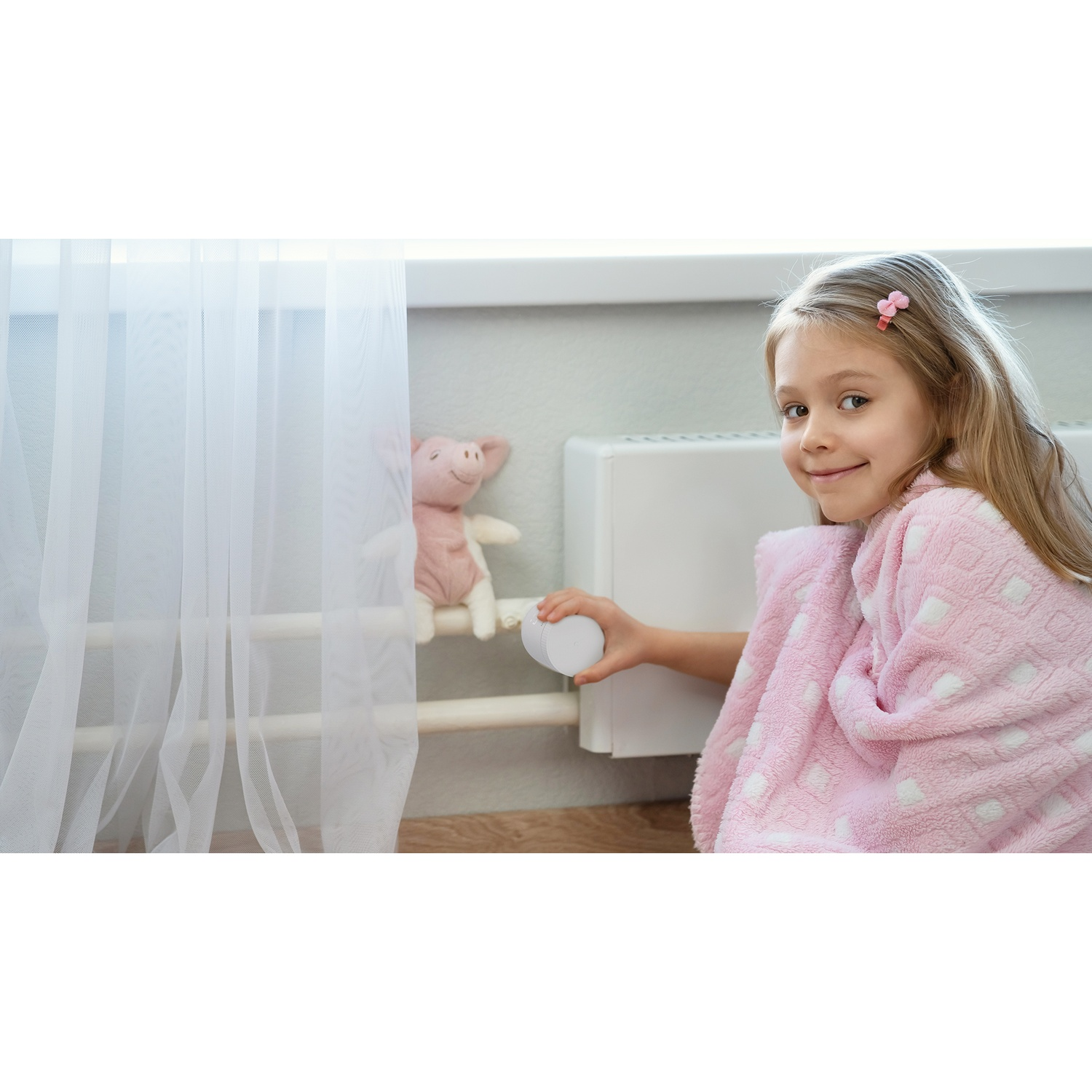 Een meisje in een roze badjas houdt een wit voorwerp vast bij een radiator. Een varkentje knuffel zit op de radiator. Wit gordijn links.