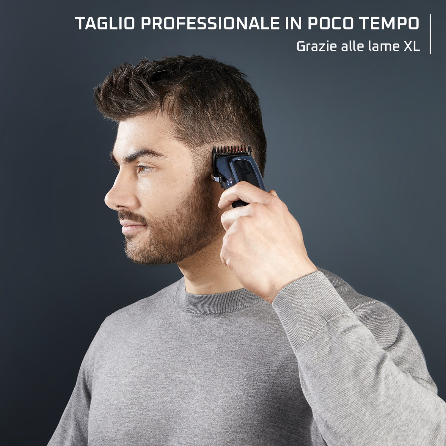 Un uomo usa un tagliacapelli per tagliarsi i capelli. Indossa un maglione grigio. Lo sfondo è grigio scuro.