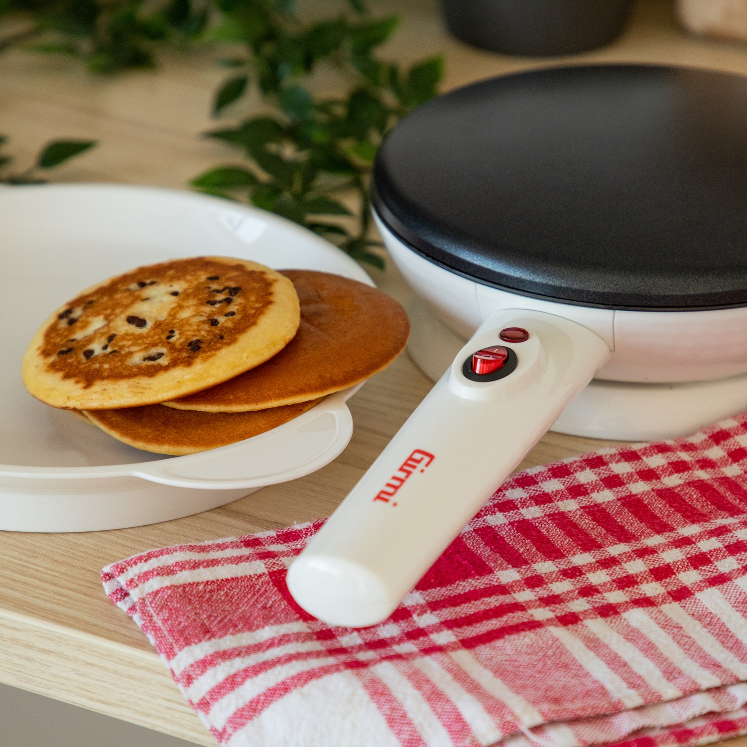 Frittelle su un piatto, crepiera e panno a scacchi rosso e bianco su una superficie di legno. Foglie verdi sullo sfondo.
