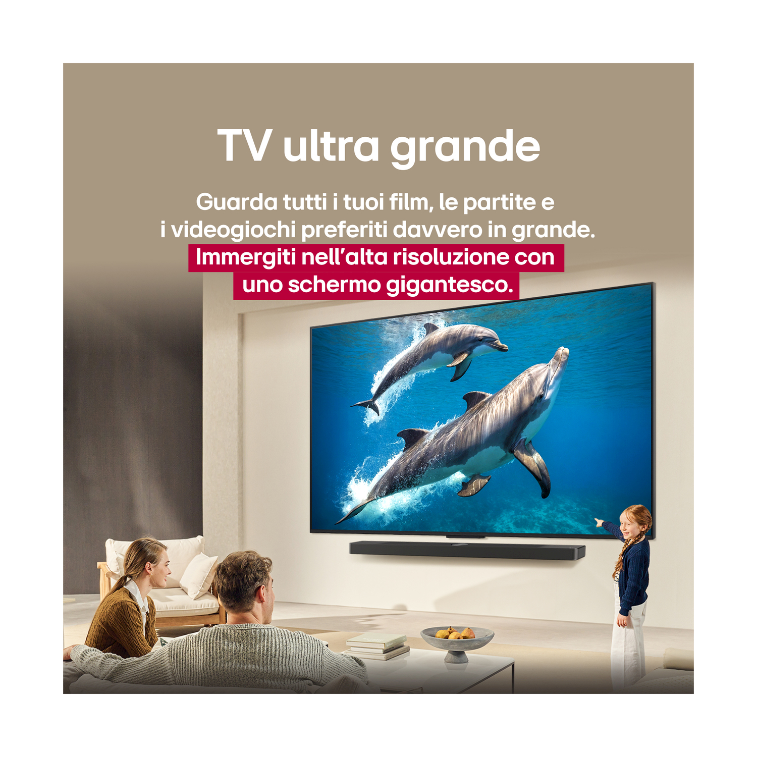 Una famiglia guarda una grande TV che mostra delfini. Una bambina indica la TV. La stanza ha un divano, un tavolino e altoparlanti.