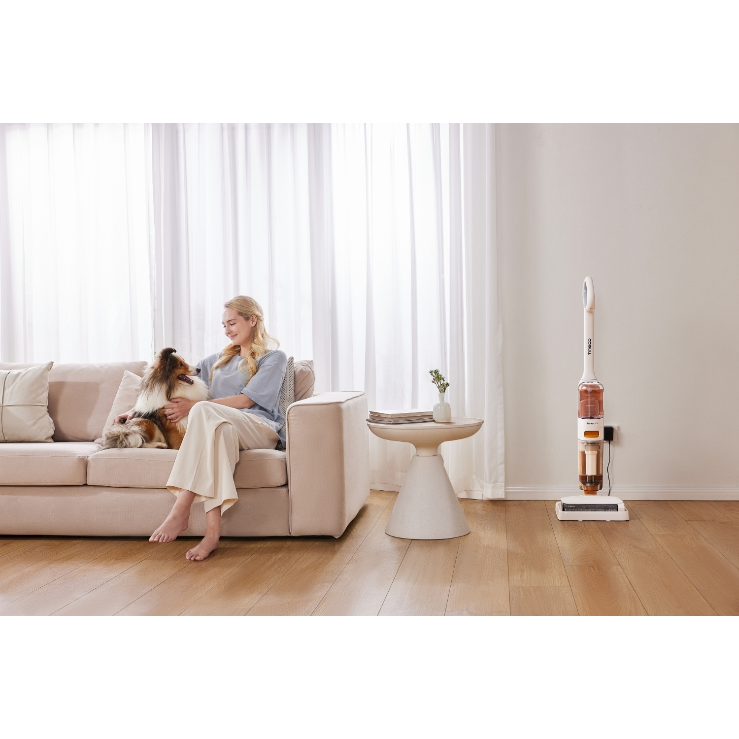 Una donna è seduta su un divano beige, che accarezza un cane. Un aspirapolvere bianco è vicino. Un tavolino ha un vaso e dei libri.