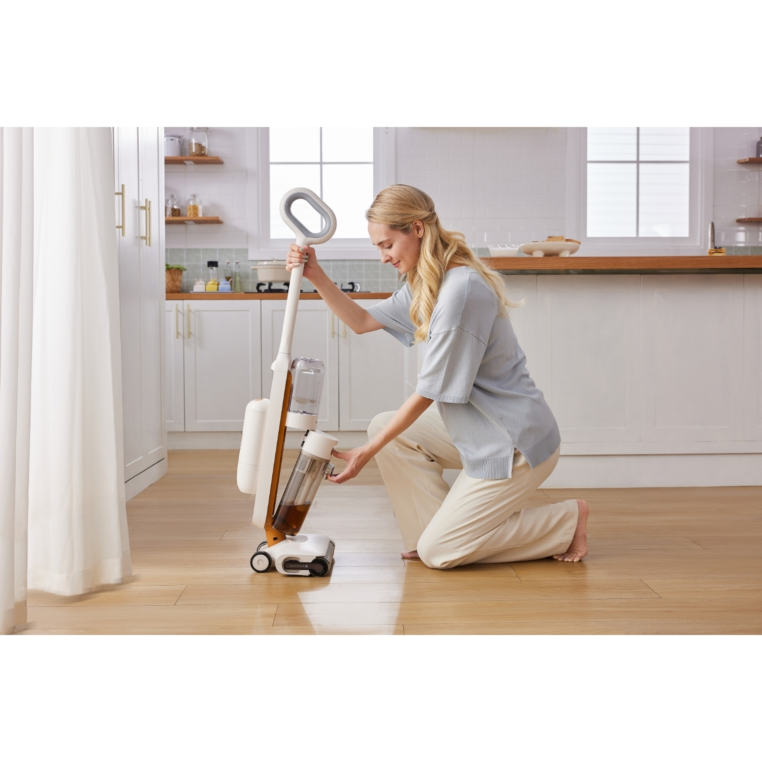 Donna che usa un pulitore per pavimenti bianco e arancione. Si inginocchia, tenendo il manico. Pavimento in legno chiaro, armadi bianchi e finestra sullo sfondo.