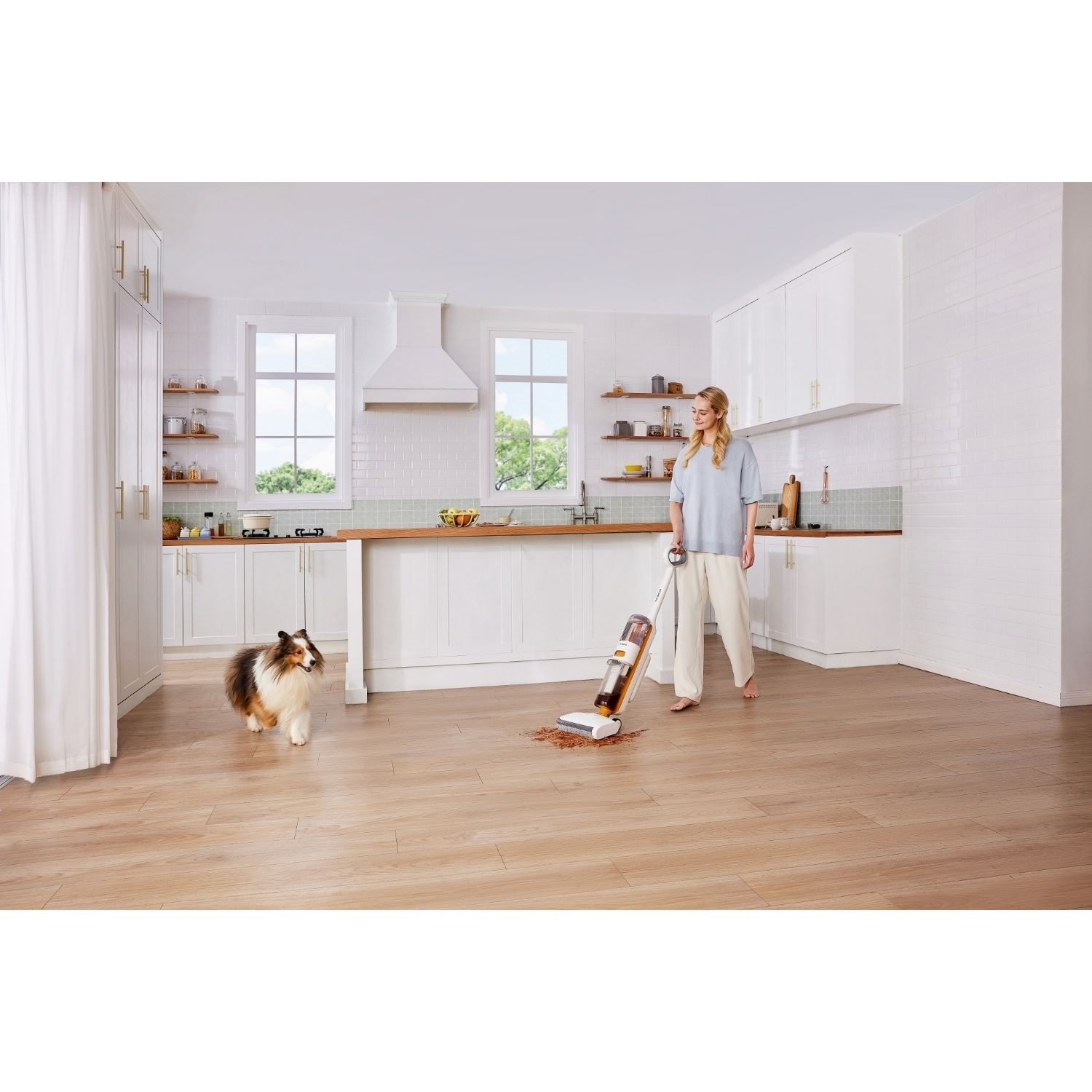 Una donna aspira il pavimento della cucina. Un cane le si avvicina. La cucina è bianca con pavimenti e armadi in legno.