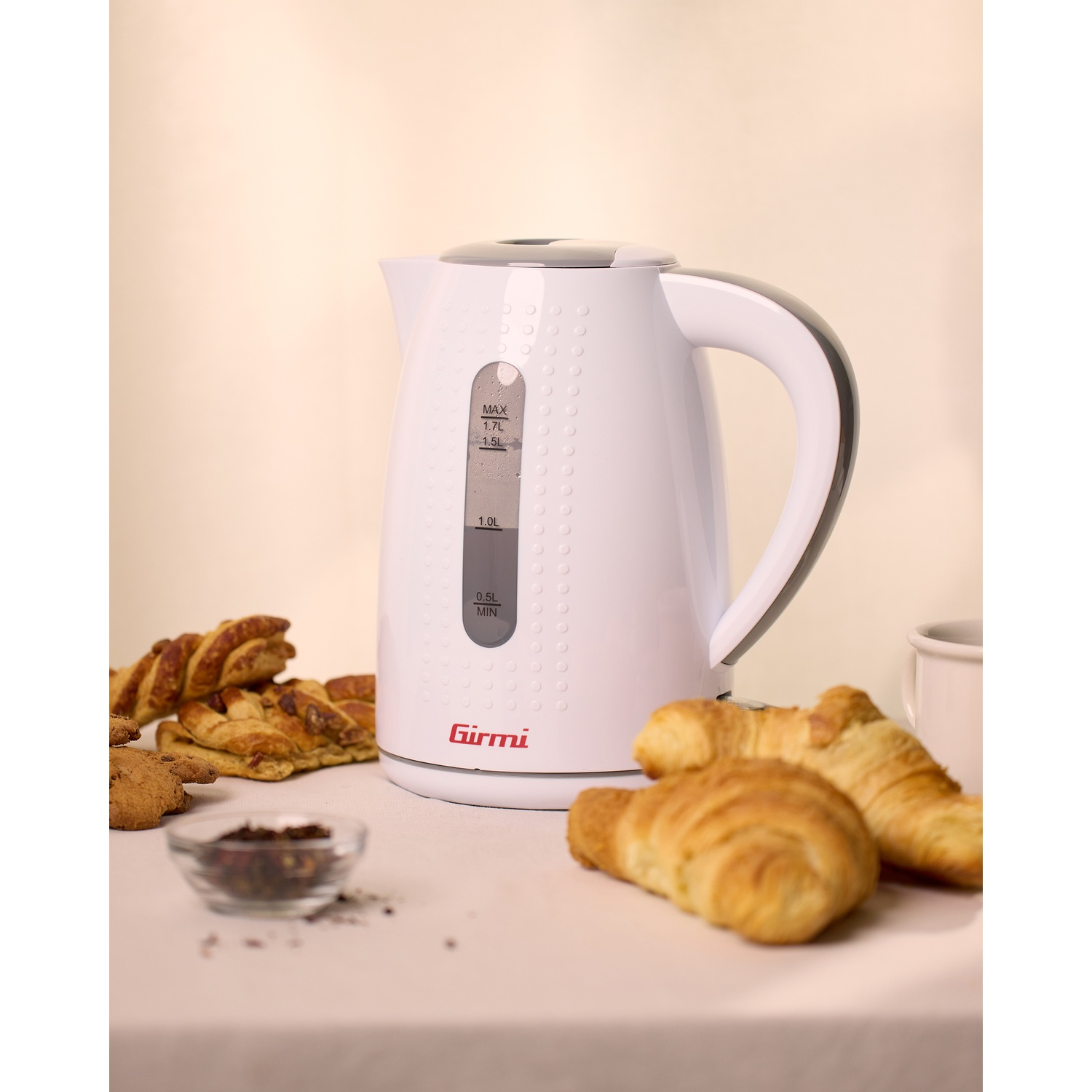 Un bollitore elettrico bianco con pasticcini e una tazza. Croissant e pane sono sul tavolo. Una piccola ciotola con contenuto scuro.