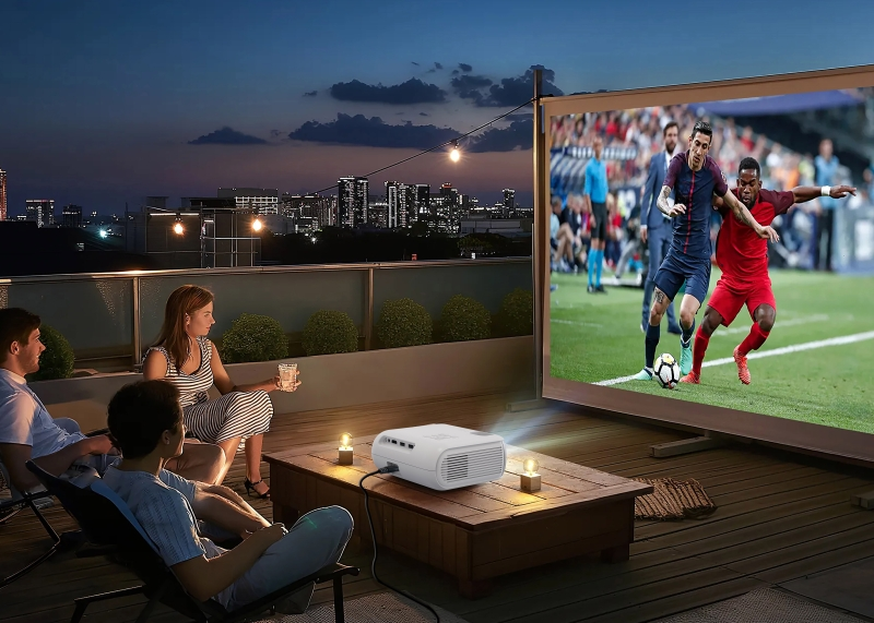 Vier Personen schauen ein Fußballspiel auf einer Leinwand. Ein Projektor steht auf einem Tisch. Stadtlichter im Hintergrund.