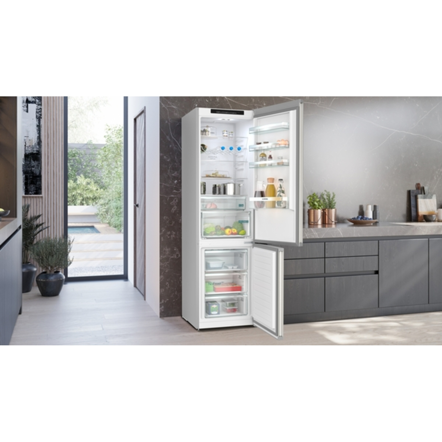 Een moderne keuken met een open koelkast. De koelkast is wit en gevuld met voedsel. De keuken heeft grijze kasten en een raam naar de tuin.