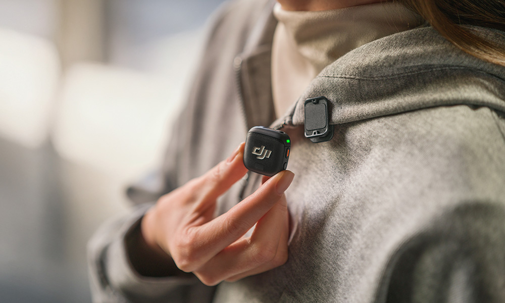 Osoba trzyma czarny mikrofon DJI. Jest przypięty do szarej bluzy z kapturem. Drugi klips jest na bluzie.