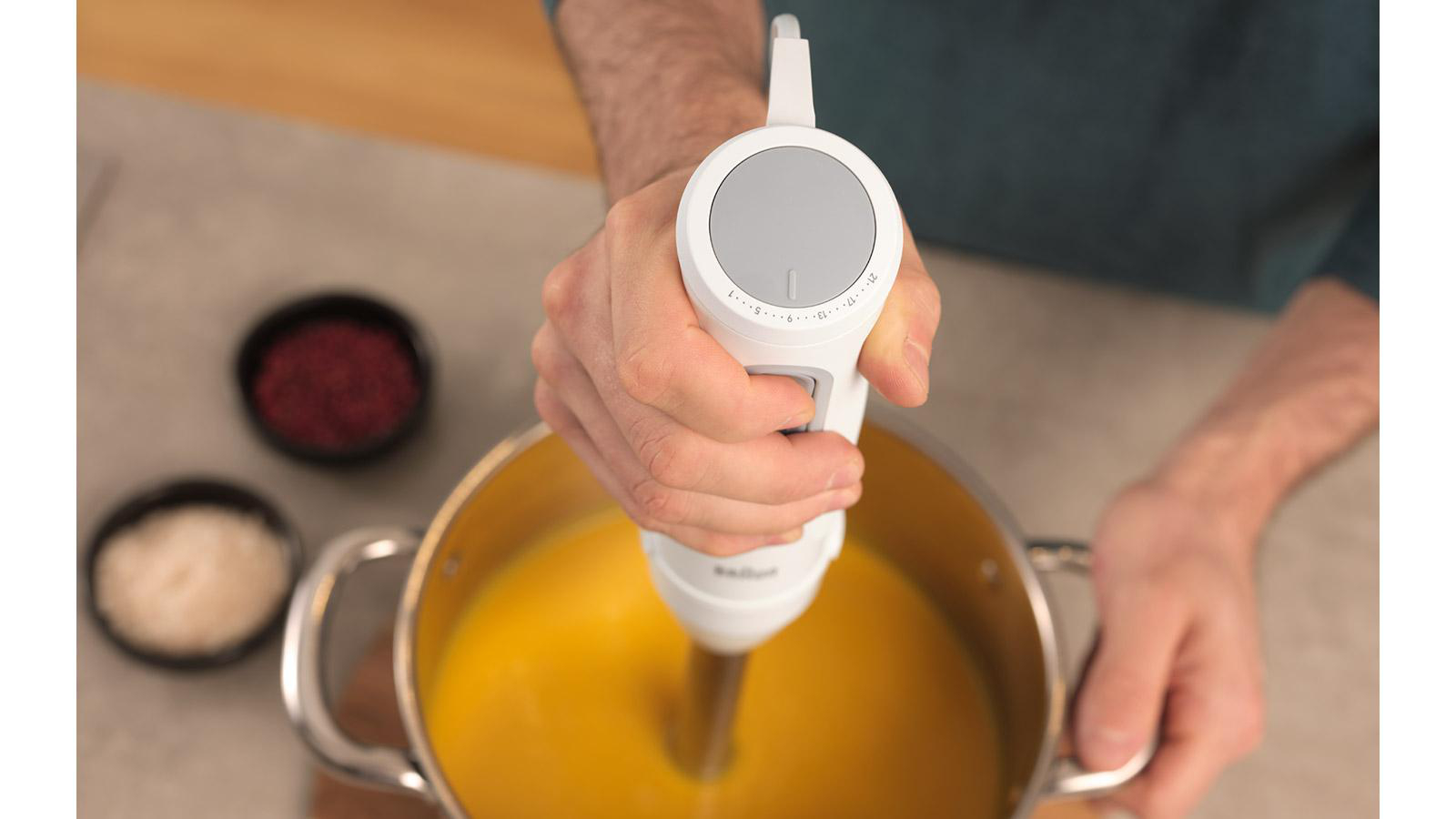 Eine Hand hält einen weißen Stabmixer über einem Topf mit orangener Suppe. Kleine Schalen mit roten und weißen Zutaten befinden sich im Hintergrund.