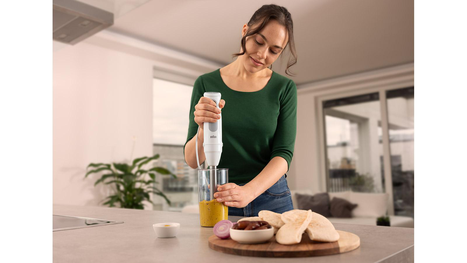 Eine Frau in grünem Oberteil benutzt einen Stabmixer. Auf der Theke stehen eine Schüssel mit Essen, Brot und Zutaten. Küchenhintergrund.