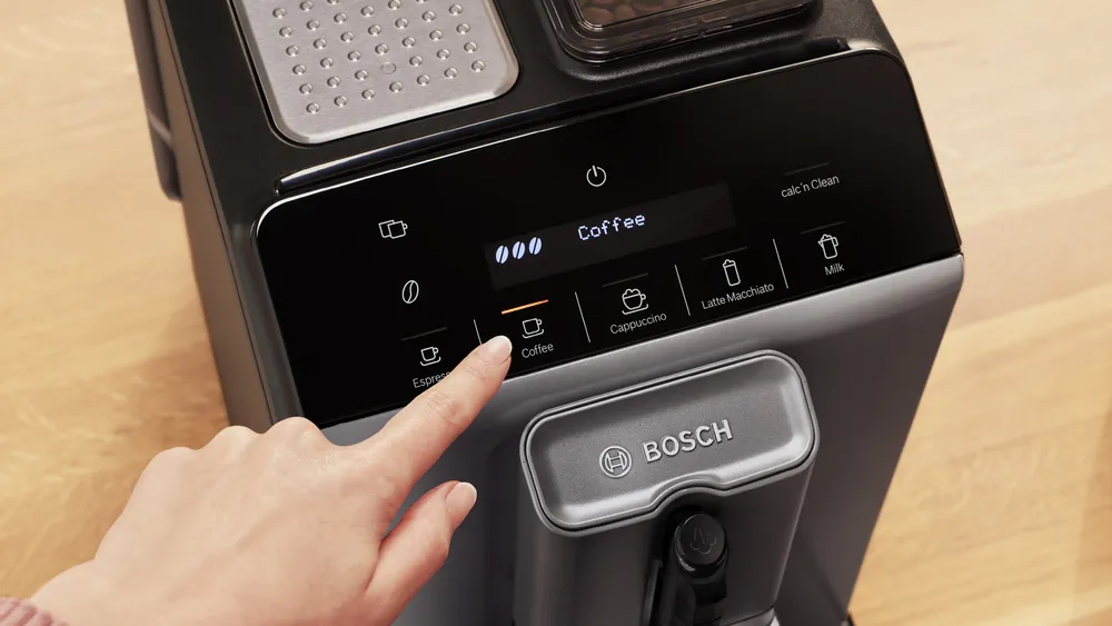 Egy kéz megnyomja a kávé gombot egy fekete Bosch kávéfőzőn. A kijelzőn a 'Coffee' felirat látható. A gép szürke és fekete.