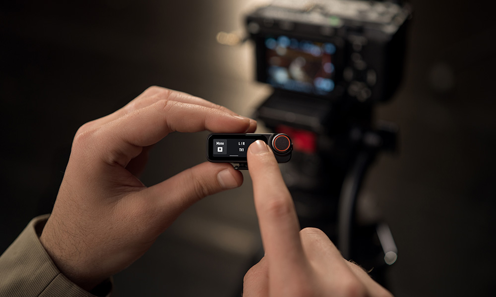 Ręce trzymające czarne urządzenie audio z ekranem, wybierające opcje. W tle aparat fotograficzny.