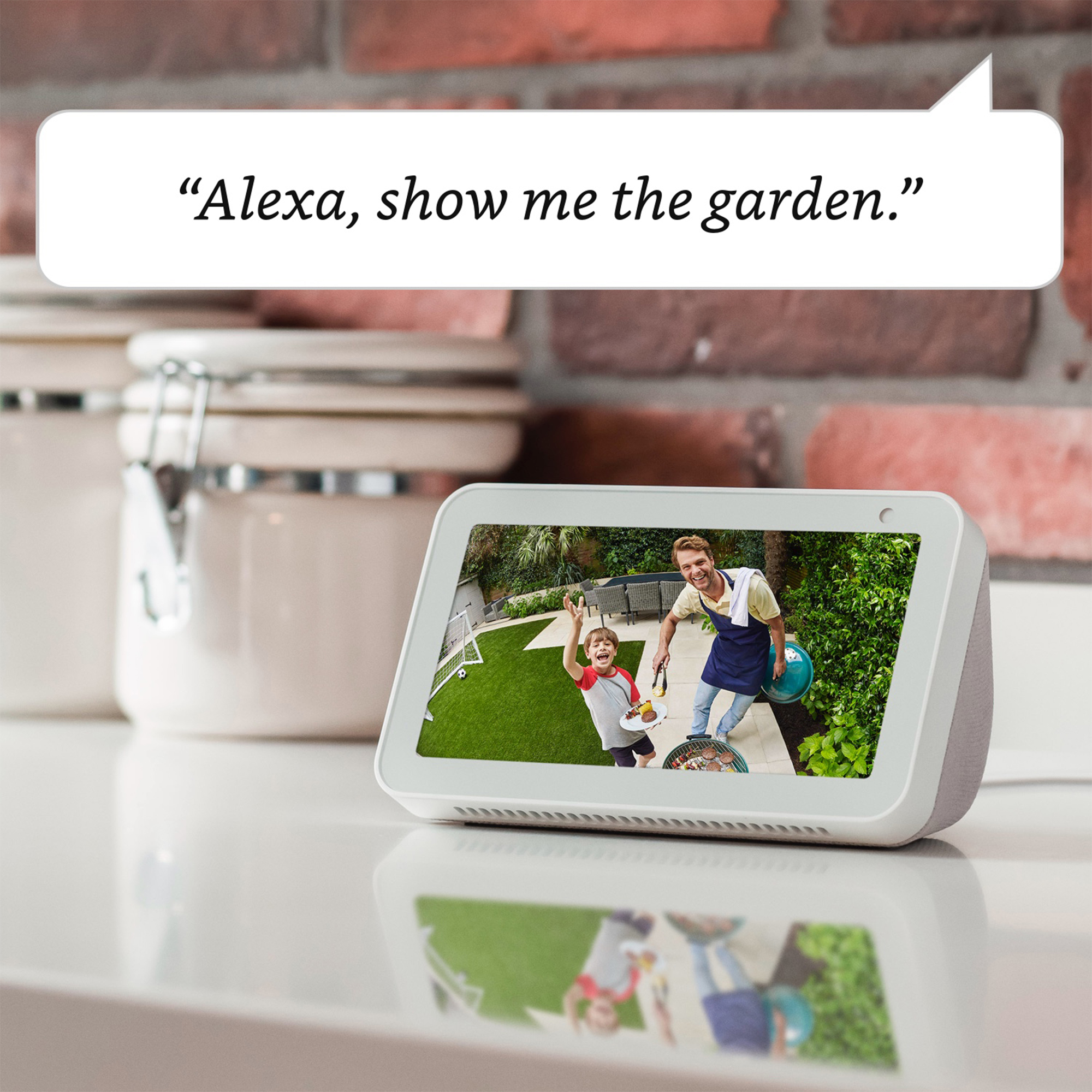 Una pantalla Echo muestra una escena de jardín con un hombre, un niño y una barbacoa. Un bocadillo dice: "Alexa, muéstrame el jardín".