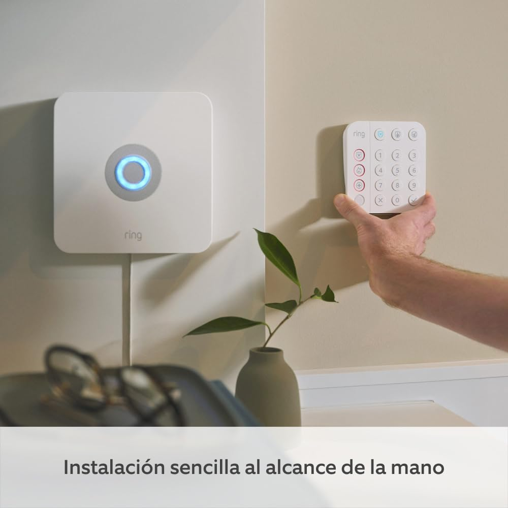 Una persona sostiene un teclado Ring junto a un panel de seguridad blanco en una pared beige. Un jarrón con una planta y gafas debajo.