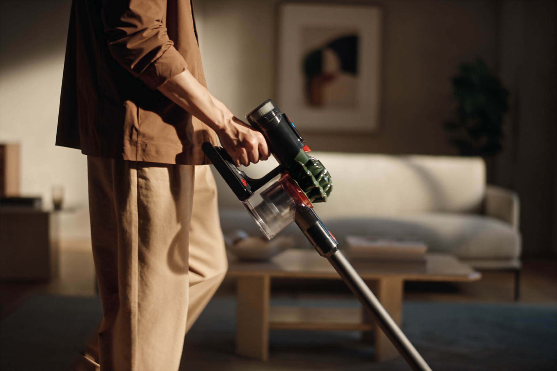 Osoba trzymająca odkurzacz. Beżowe spodnie i brązowa koszula. Tło: sofa, stolik kawowy i dzieło sztuki w salonie.