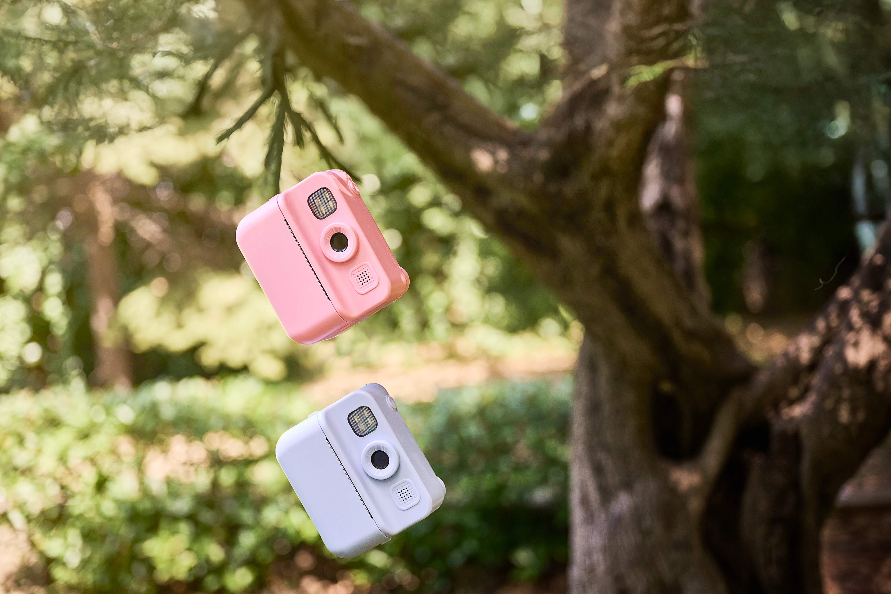 Pembe ve beyaz iki anında fotoğraf makinesi, bulanık bir ağaç ve yeşil yaprakların önünde havada duruyor.