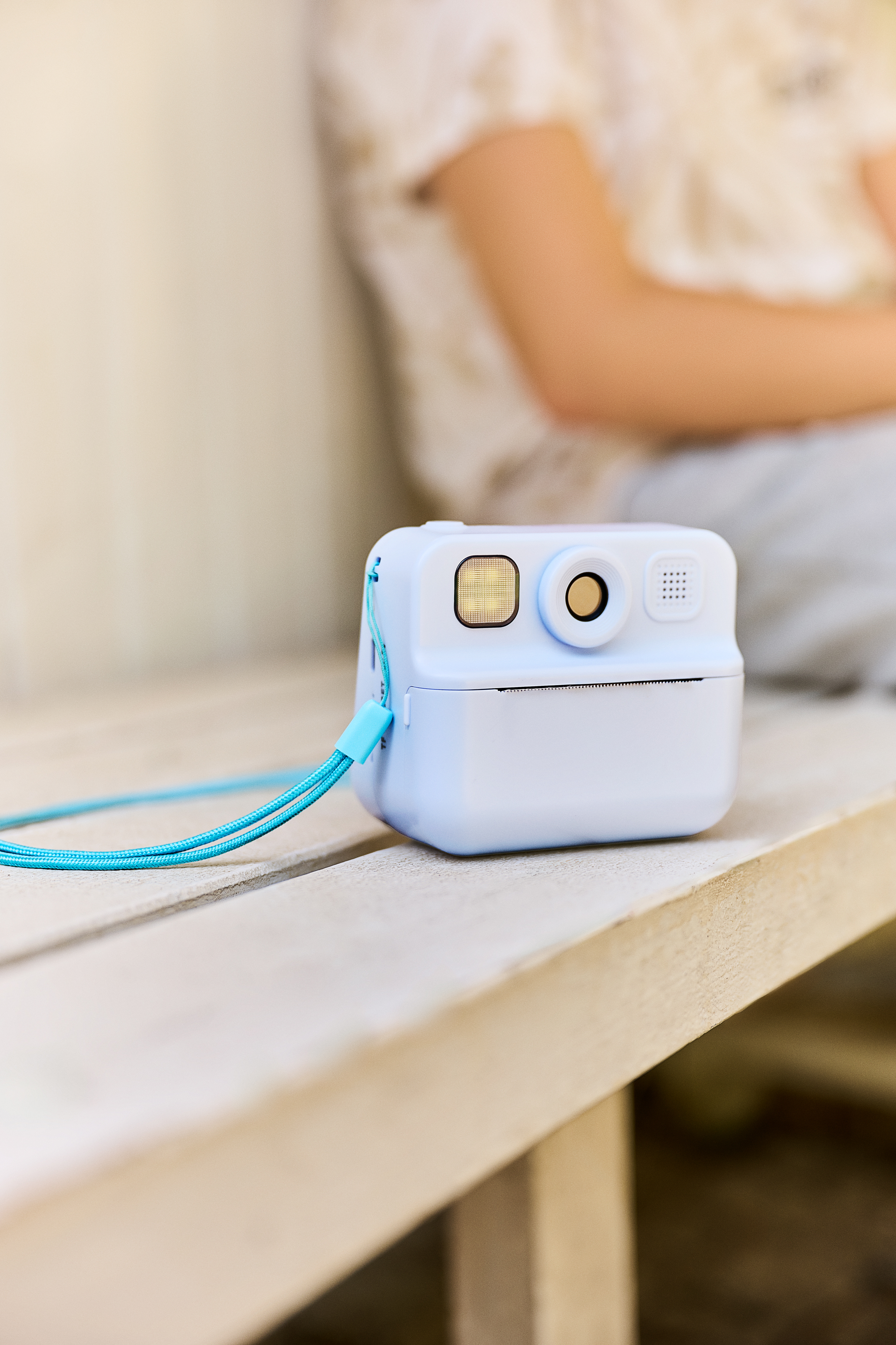 Açık mavi bir anlık fotoğraf makinesi ahşap bir bankta duruyor. Arka planda bulanık bir kişi var.