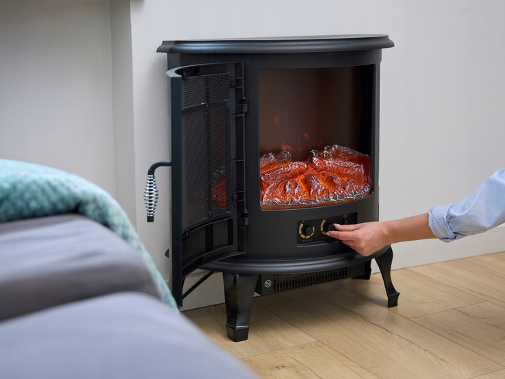 Otwarty czarny kominek elektryczny. Ręka obraca pokrętło. Sztuczne płomienie świecą na pomarańczowo. Na pierwszym planie łóżko.