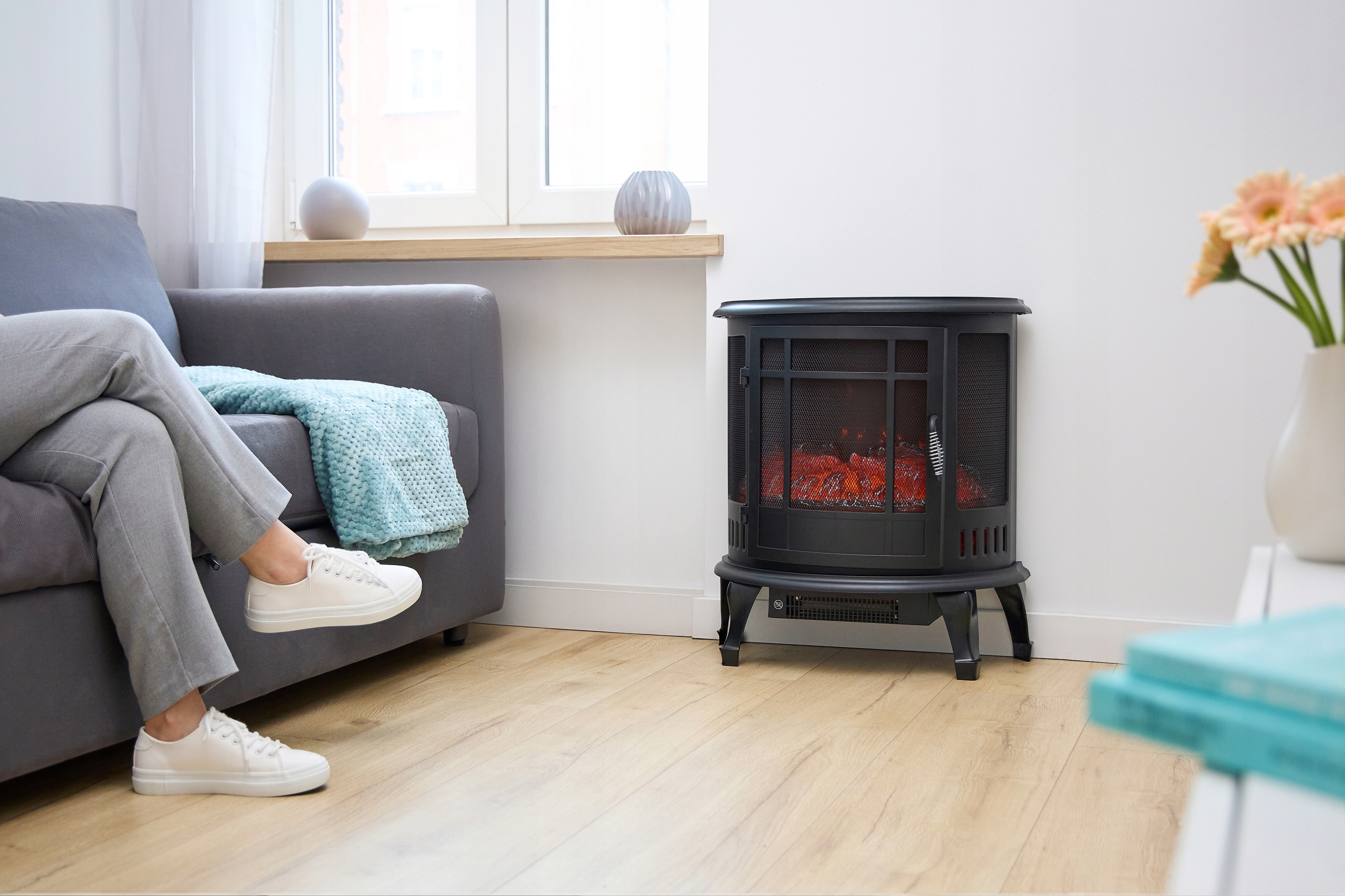 Osoba siedzi na szarej sofie w pobliżu czarnego kominka. W pobliżu jest koc i kwiaty. Podłoga jest drewniana.