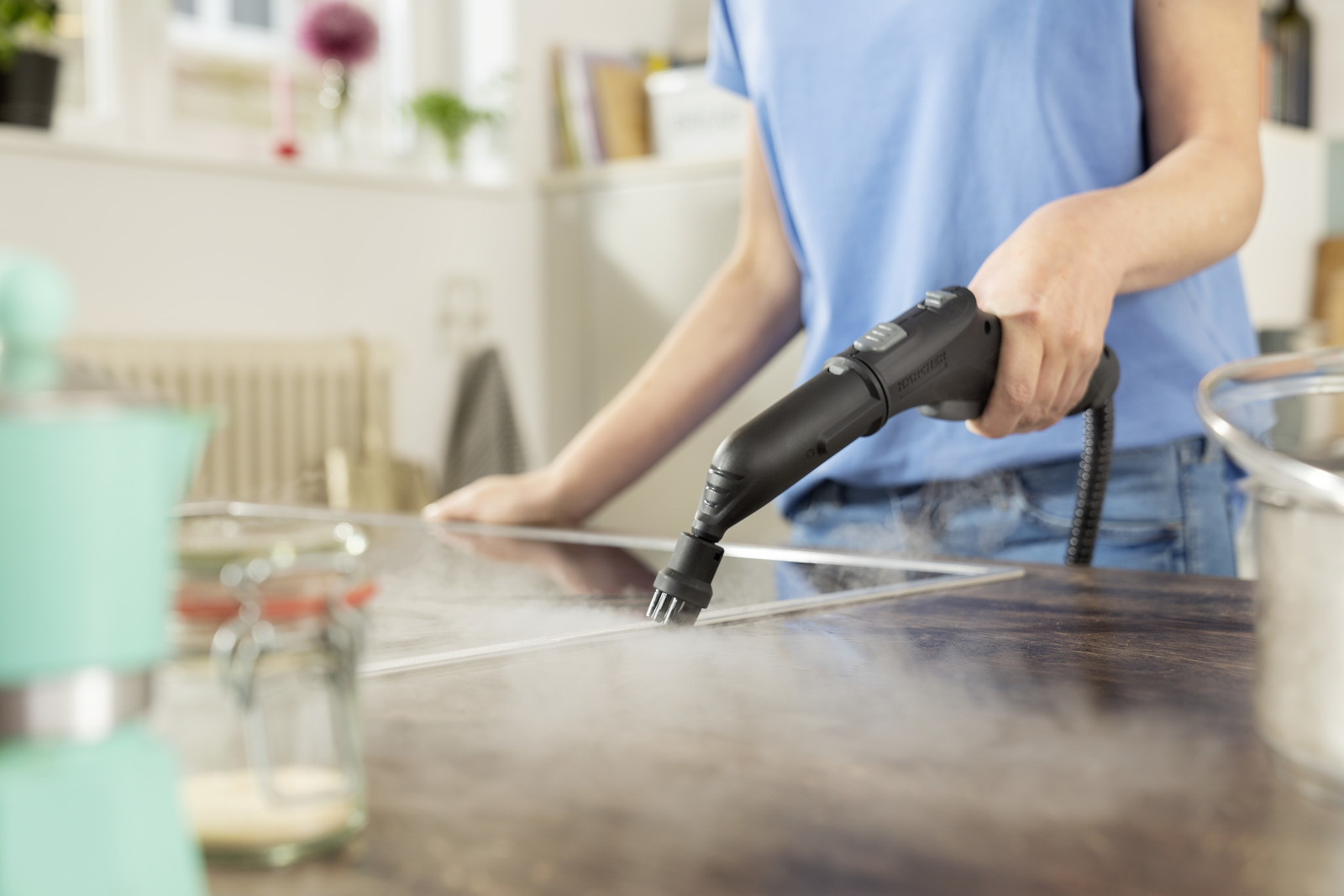 Osoba czyści drewnianą powierzchnię za pomocą parownicy. Biała para unosi się. Tło kuchni z garnkiem, słoikami i turkusowym ekspresem do kawy.