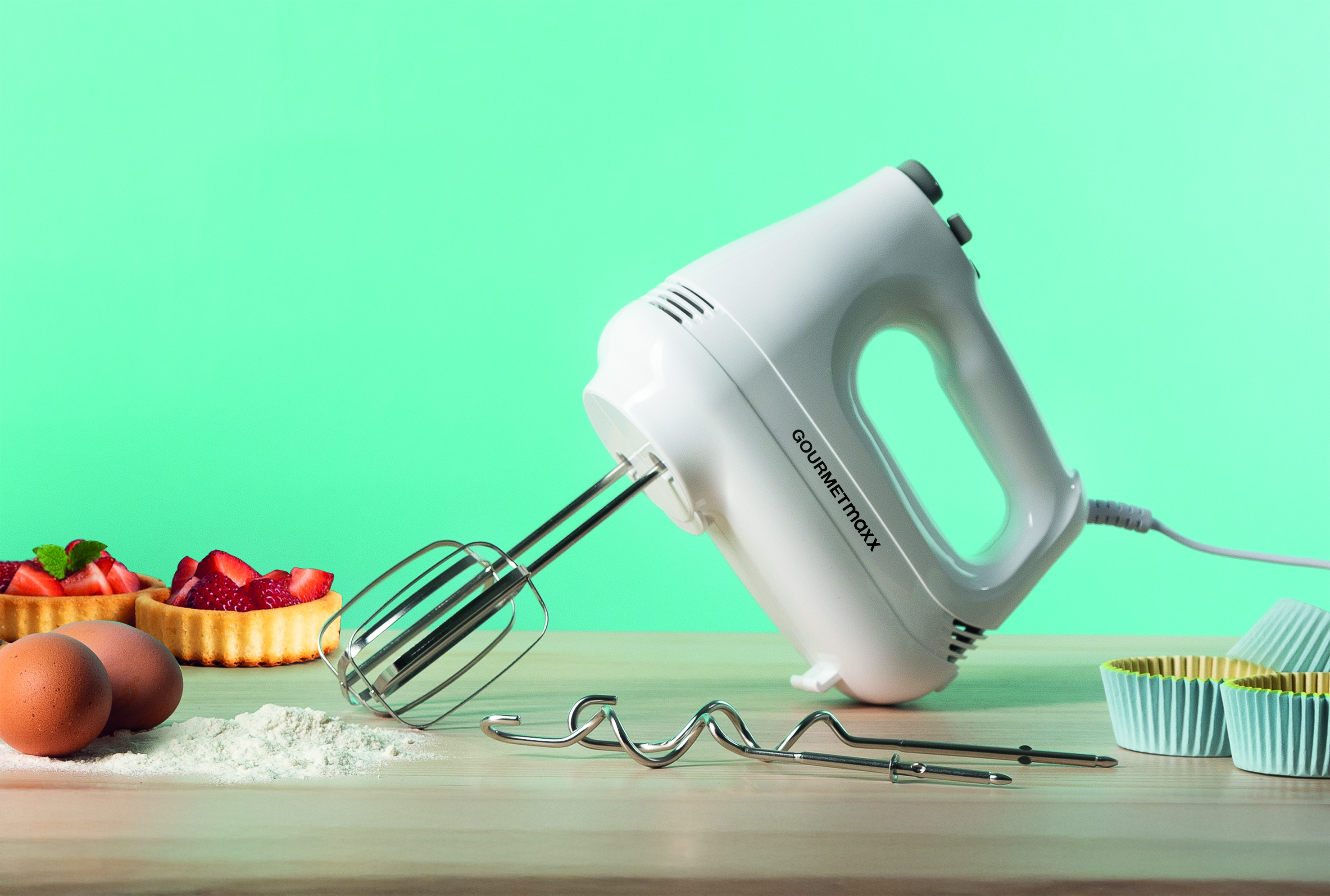 Weißer Handmixer mit Schneebesen auf Holz. Erdbeertörtchen, Eier und Mehl sind im Bild.