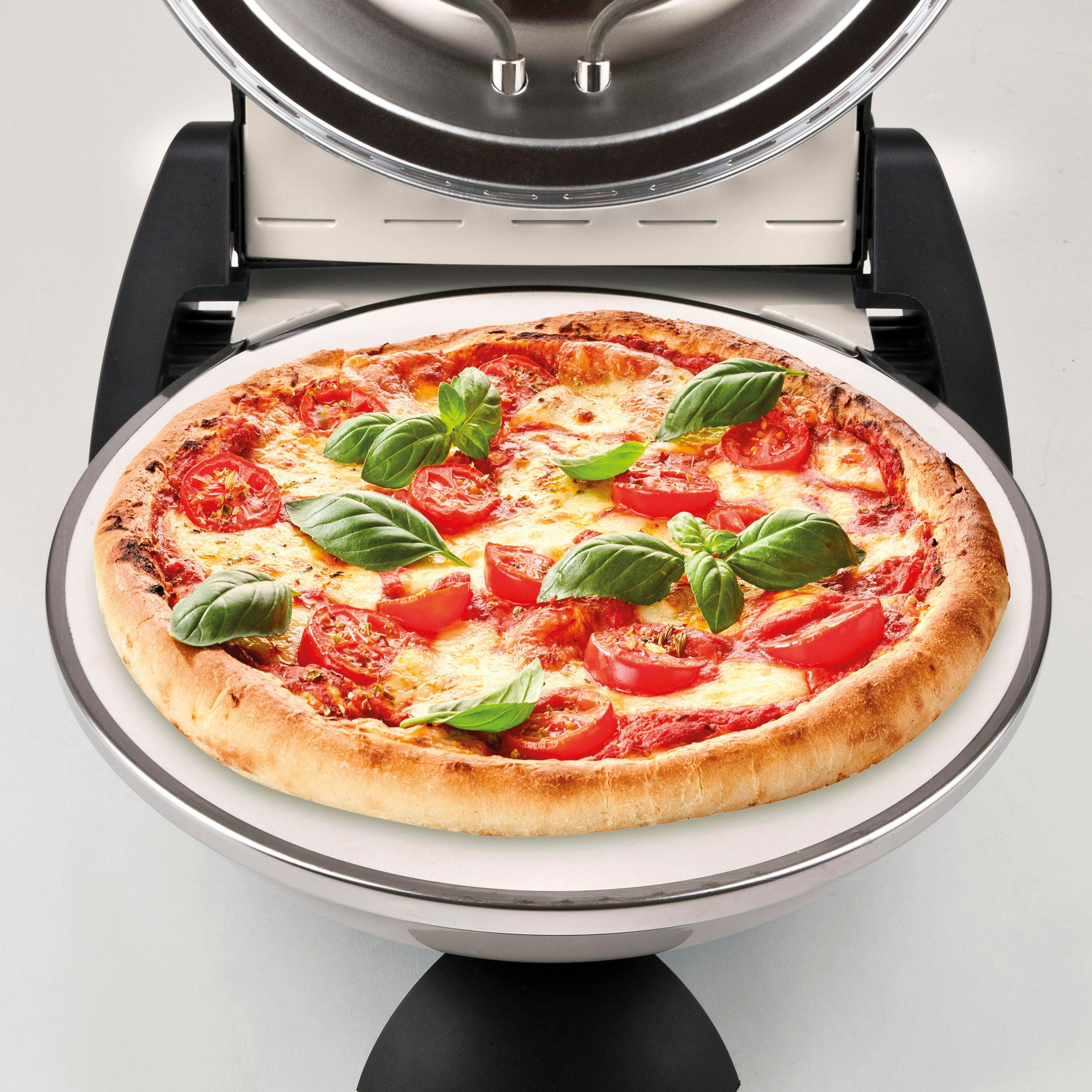 Eine Pizza mit Tomaten und Basilikum auf einem weißen Teller in einem Pizzaofen. Der Ofen ist silber und schwarz.