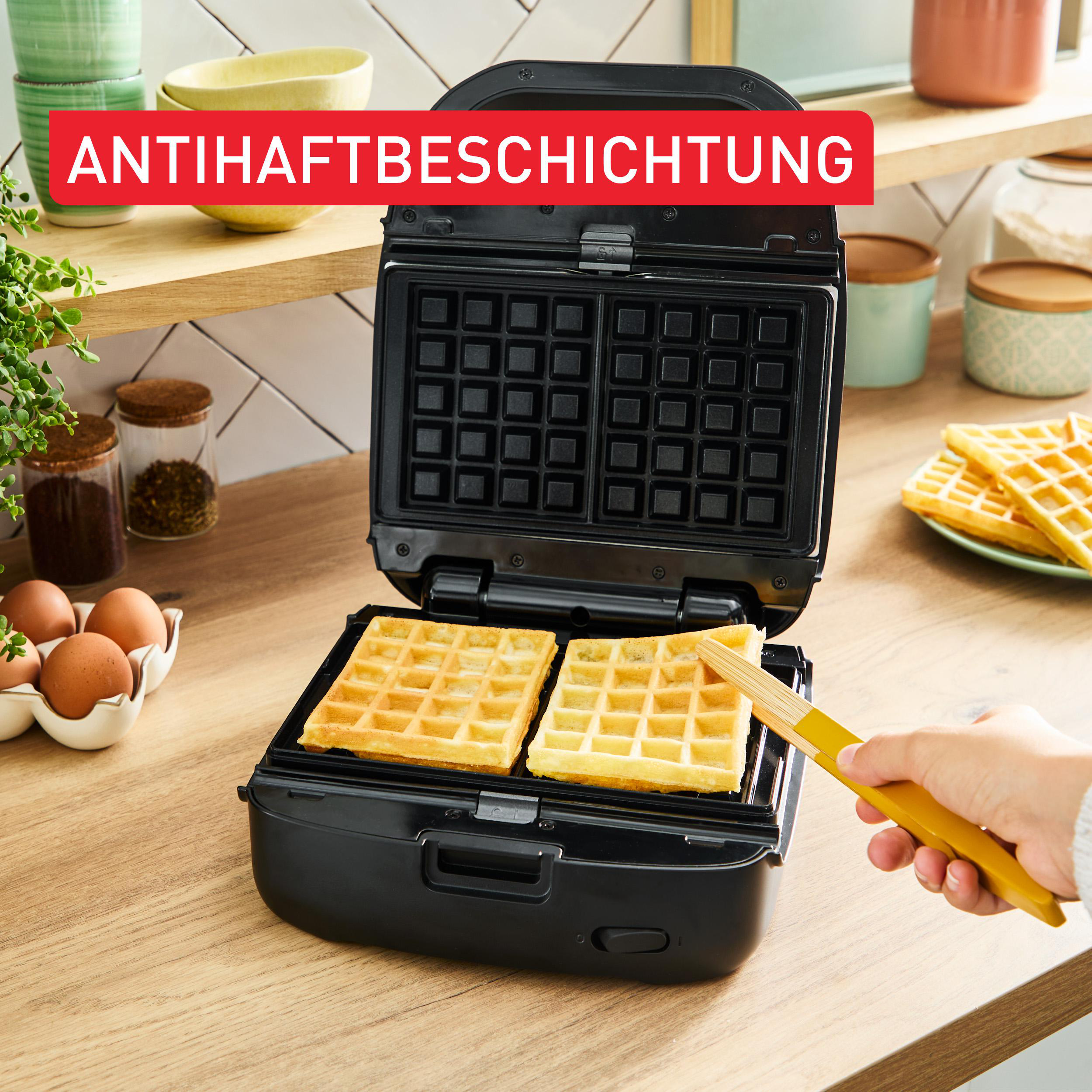 Ein Waffeleisen ist geöffnet und zeigt zwei fertige Waffeln. Eine Hand hält einen gelben Spatel, bereit, eine Waffel zu entnehmen.