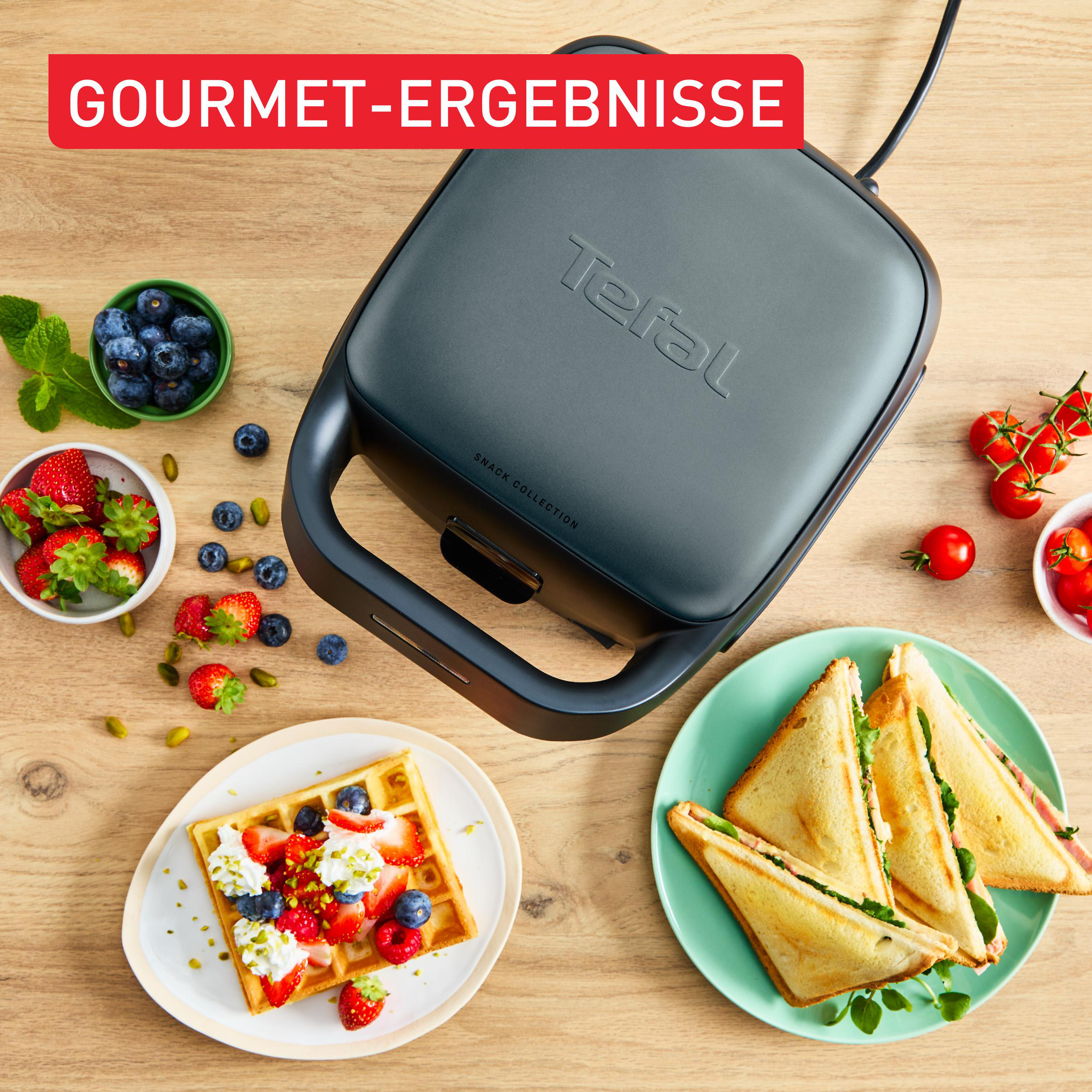 Ein Waffeleisen mit Snacks. Waffeln, Sandwiches, Beeren und Tomaten sind darum angeordnet. Die Szene befindet sich auf einem Holztisch.